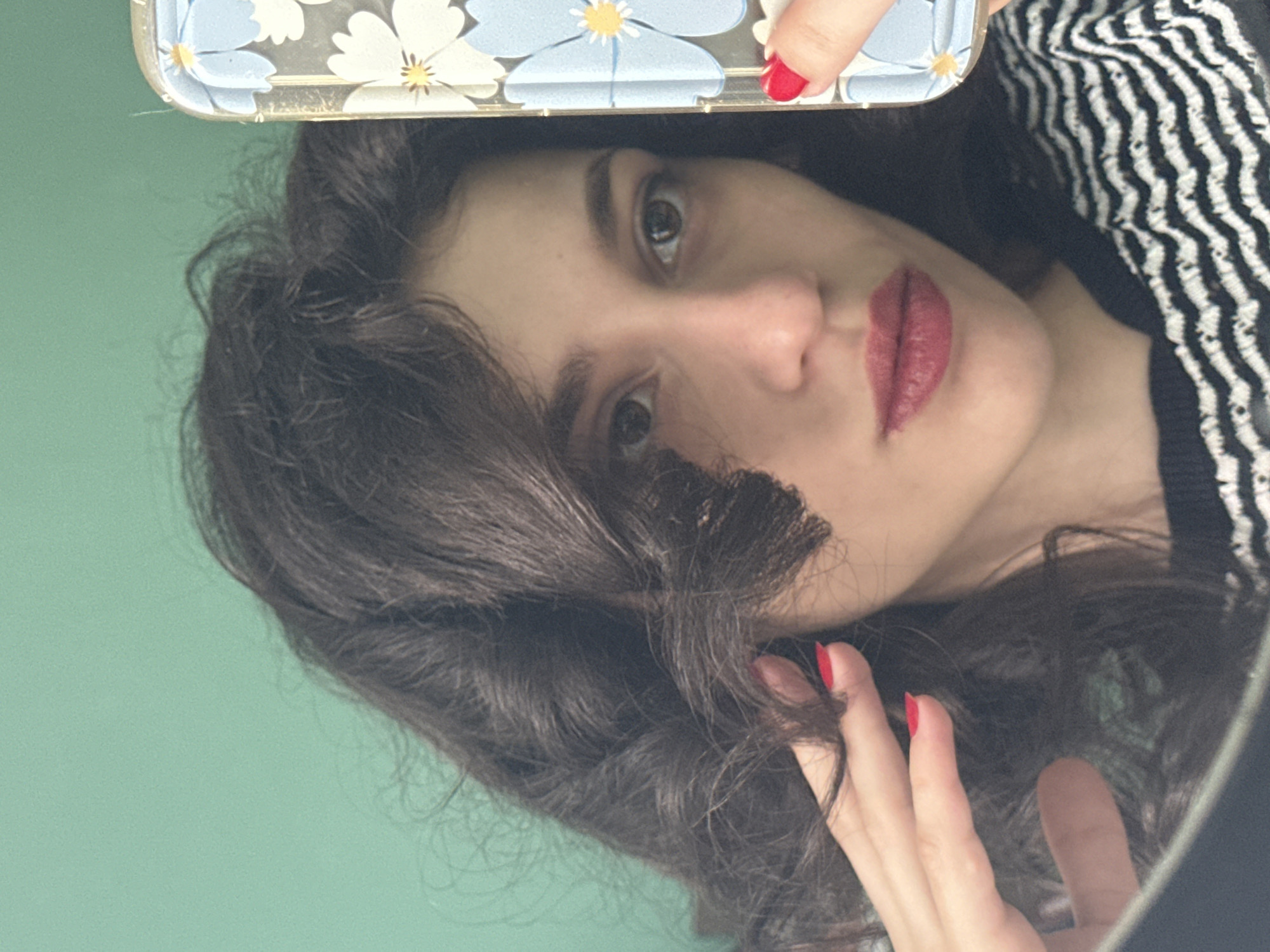 A young woman with dark, wavy hair and red nail polish is taking a selfie in front of a green wall. She is wearing a black and white striped shirt. She is holding her phone, which has a blue and white floral case, in her right hand, and touching her hair with her left hand. The image is a close-up shot, focusing on her face and upper body. There is no information about the location in Sofia, Bulgaria.