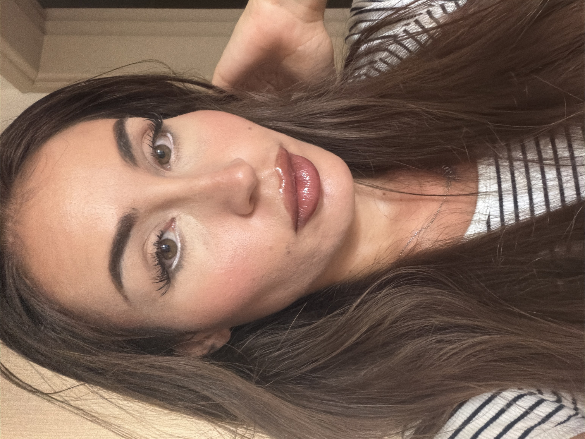 A medium shot shows a woman with long, dark brown hair and fair skin, looking directly at the camera. She has pronounced cheekbones and is wearing makeup, including white eyeliner and lip gloss. Her hair frames her face, with some strands falling over her left shoulder. She is wearing a black and white striped top, and a silver necklace is visible. Her right hand is raised and touching her head, obscuring part of her hair. The background appears to be indoors, with a neutral-colored wall and a doorframe visible to the right. The lighting is warm and diffused. There is no visible indication of the image being taken in Tersefanou, Cyprus.