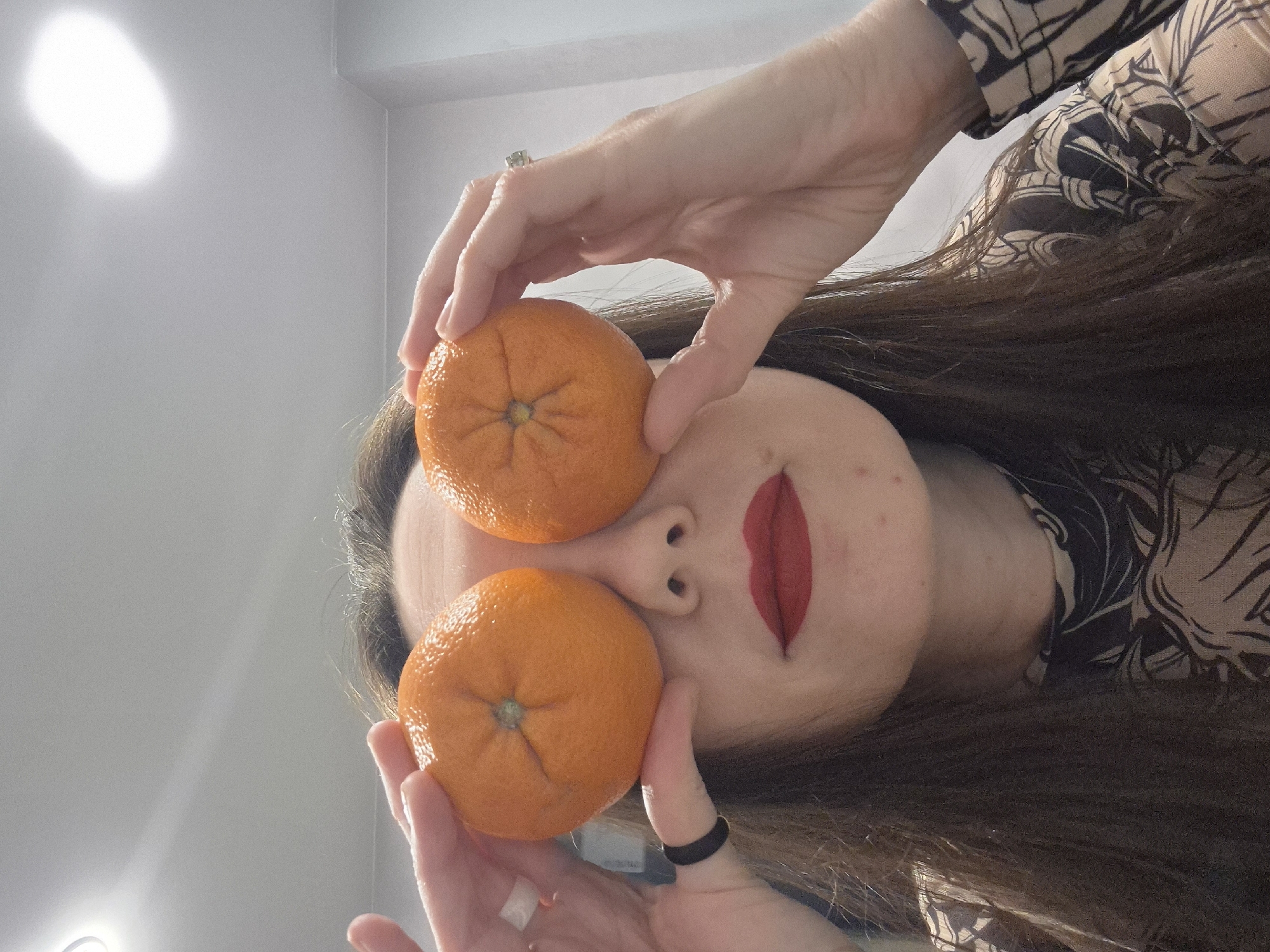 A woman with long brown hair holds two tangerines in front of her eyes. She has red lipstick on and is wearing a patterned shirt. The photo is taken from a low angle, looking up at her. The background is a light grey wall. There is no information to suggest that the image was taken in Chaikovsky, Russia.