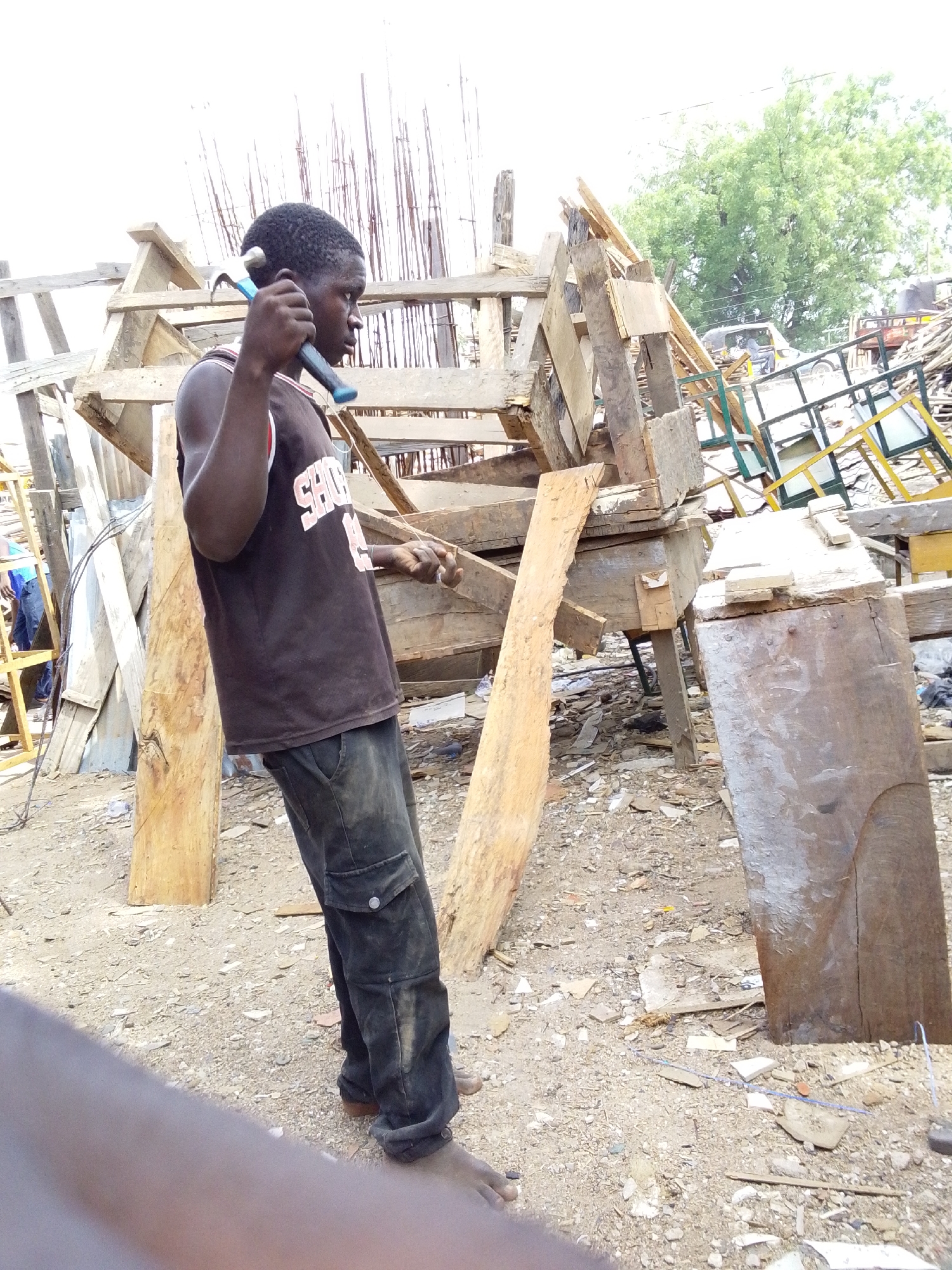 The image depicts a young man in Jimeta, Nigeria, engaged in carpentry work. He stands in a cluttered outdoor workspace filled with wood scraps and building materials. The man, wearing a dark sleeveless shirt and dark trousers, holds a hammer to his shoulder. Around him are various pieces of wood, some forming a structure, and other construction debris. The ground is dirt with scattered small wood fragments. In the background, other wooden structures and green vegetation are visible.