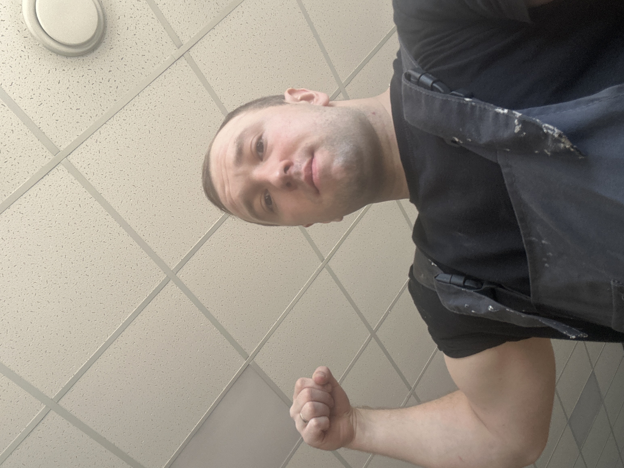 A man with a shaved head, light complexion, and wearing a dark t-shirt and dark work apron is seen in a low-angle shot indoors. He is looking directly at the camera. His right fist is clenched. Above him is a drop ceiling with recessed lighting. There is no information about the location in the city of Noviy, Russia.