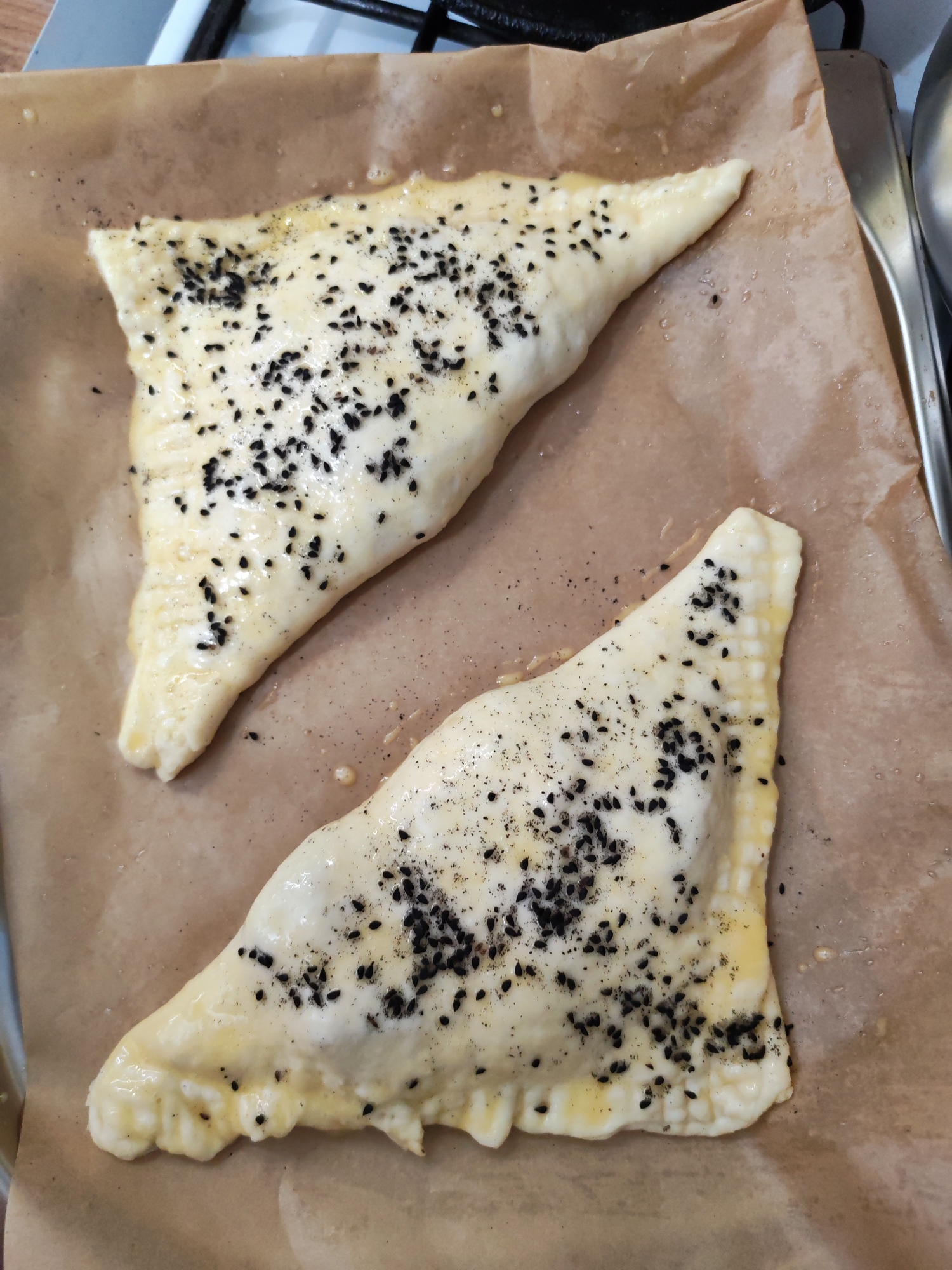 The image displays two uncooked, triangular pastries, possibly savory turnovers or samsa, resting on a sheet of brown parchment paper. The pastries are light in color, suggesting a dough such as puff pastry, and their surfaces are brushed with a glossy egg wash. They are liberally sprinkled with black sesame seeds and some finer dark specks, possibly ground pepper or other spices. The edges of both pastries are clearly crimped, indicating they have been sealed, likely to contain a filling.
The parchment paper, which covers a baking sheet or tray, shows some glistening marks, likely from oil or the egg wash. In the upper left background, part of a white and black stove grate is visible, suggesting a kitchen environment. On the right side, a metallic edge of what appears to be another baking pan or kitchen utensil can be seen. The overall lighting is bright and even, typical of an indoor setting.
No people are visible in the frame, and there is no discernible text. The scene depicts food prepared and ready for baking. While the location context of Izhevsk, Russia, is provided, there are no specific visual cues within the image itself to confirm this geographical detail.