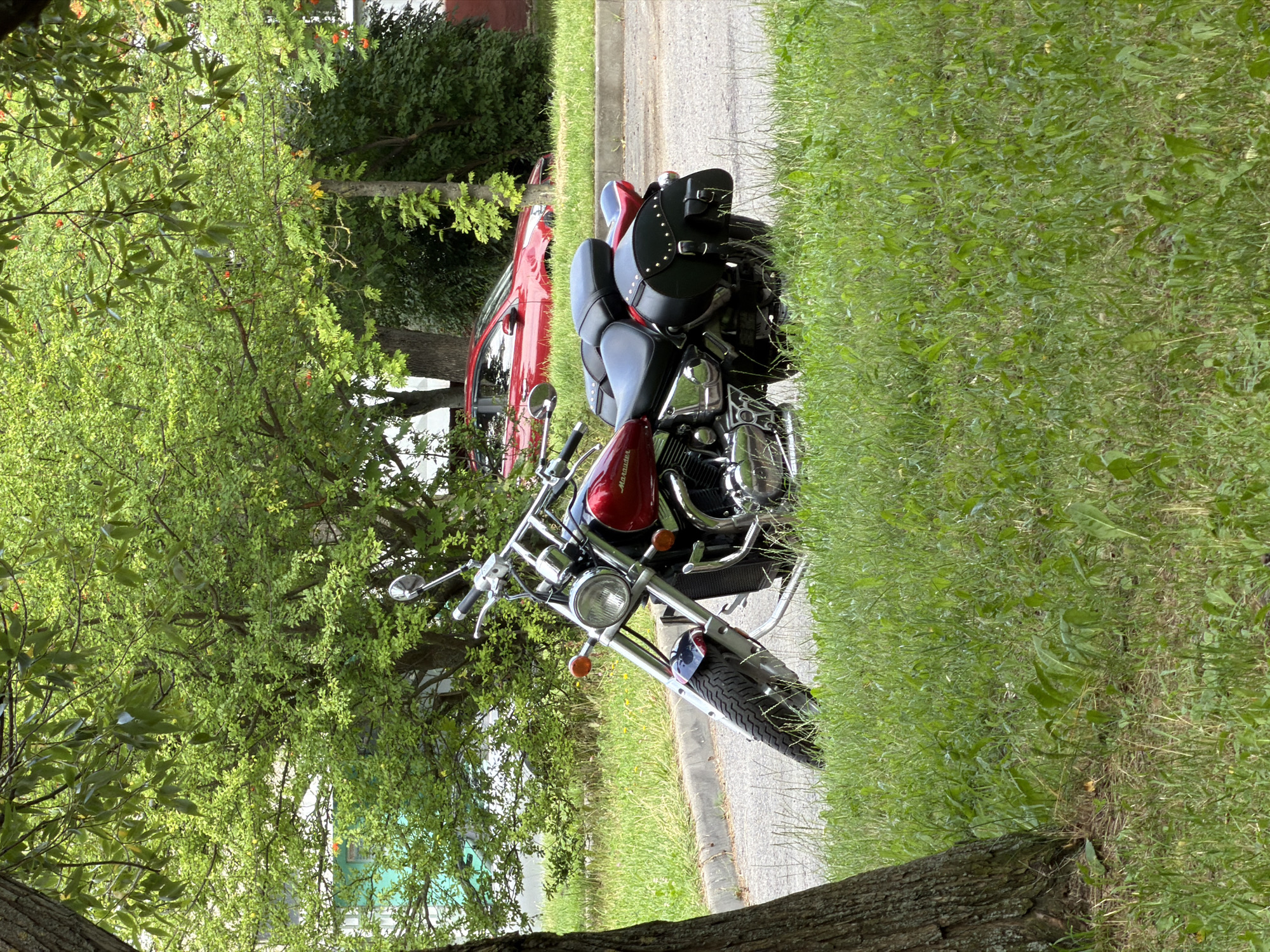 A red and black cruiser motorcycle is parked on the side of a paved road, with its front wheel turned to the left, resting on the grass verge. The motorcycle has leather saddlebags on the rear. In the background, a red car is parked further down the road, partially obscured by trees and foliage. The scene appears to be in a residential area, with green grass and leafy trees. The location is identified as Minsk, Belarus.
