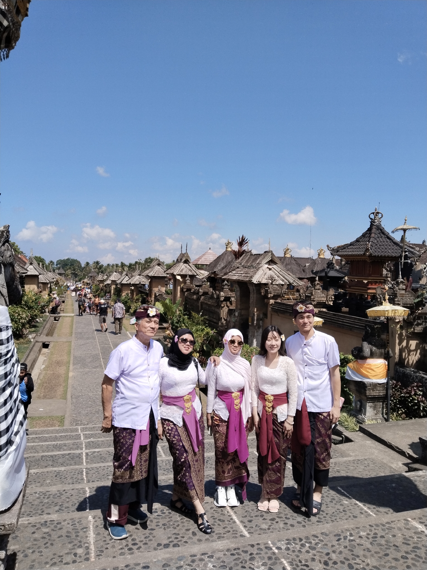 In the city of Bangli, Indonesia, five individuals, three women and two men, are posing for a photograph on a stone staircase. The individuals are dressed in traditional Balinese attire, consisting of sarongs, blouses, and head coverings. The two men are wearing white shirts and headbands, while the women are wearing intricately patterned sarongs and tops, with two of them also wearing headscarves. They are standing in front of a row of traditional Balinese buildings with tiered roofs, surrounded by lush vegetation. The sky is clear and blue, with a few scattered clouds. In the background, other tourists can be seen walking along a pathway, suggesting this is a popular tourist destination.