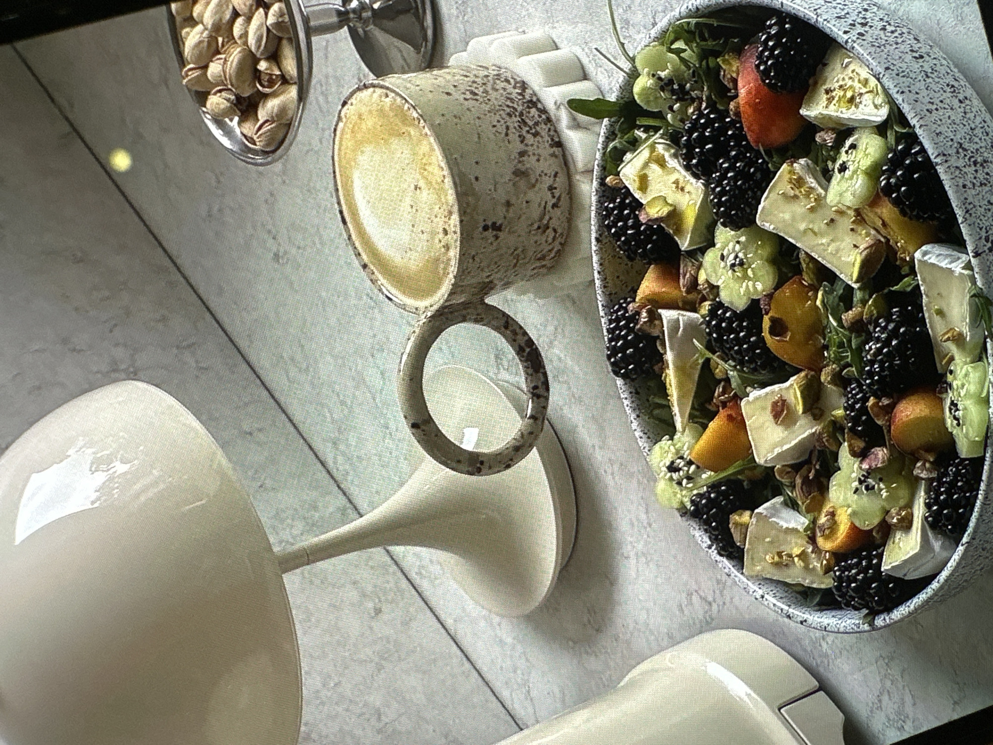 The scene presents a still life arrangement viewed from above. A bowl, featuring a grey speckled exterior, contains a salad composed of blackberries, peaches, cheese (appearing to be brie or a similar soft cheese), and green leafy vegetables. Adjacent to the bowl is a ceramic mug with a speckled, earthy tone, filled with a pale liquid. To the left, a stemware piece with a wide, opaque bowl sits near a smaller dish filled with pistachios. The background appears to be a light-colored, possibly marble, surface. There is no indication as to the location or any mention of Yerevan, Armenia in the image. Nor are there any people evident in the photo.