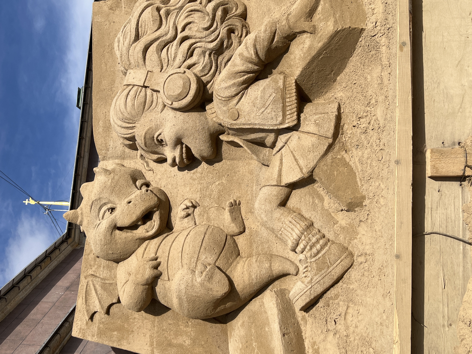 This media file captures a vibrant and detailed relief sculpture depicting a playful interaction between a young girl and a friendly creature, set against a clear blue sky in Saint Petersburg, Russia. The sculpture is crafted from a light brown, sandy material, giving it a textured appearance.
The main subjects are two sculpted figures. On the upper right, a girl with long, wavy hair and large headphones grins widely, leaning forward with her body angled. She wears a jacket with visible details like buttons and a pocket. Below her, a small, cartoon-like dragon or mythical creature lies on its back, also smiling cheerfully with wide eyes. Its body shows ribbed textures, and it has small wings on its back. Both figures exude an emotion of joy and camaraderie, suggesting a lighthearted moment between companions.
The activity taking place is a depiction of playful interaction, likely part of a public art installation or sand sculpture festival. The meticulous detailing on the hair, clothing, and creature's features indicates skilled craftsmanship.
Notable details include the bright, clear blue sky with a few wispy clouds, indicating sunny weather. The strong shadows cast by the sculpture suggest it is mid-day or afternoon. To the left, a building's roofline and a power line are visible, while to the right, a wooden structure, possibly scaffolding or part of a temporary display, frames the sculpture. No visible text appears in the image.