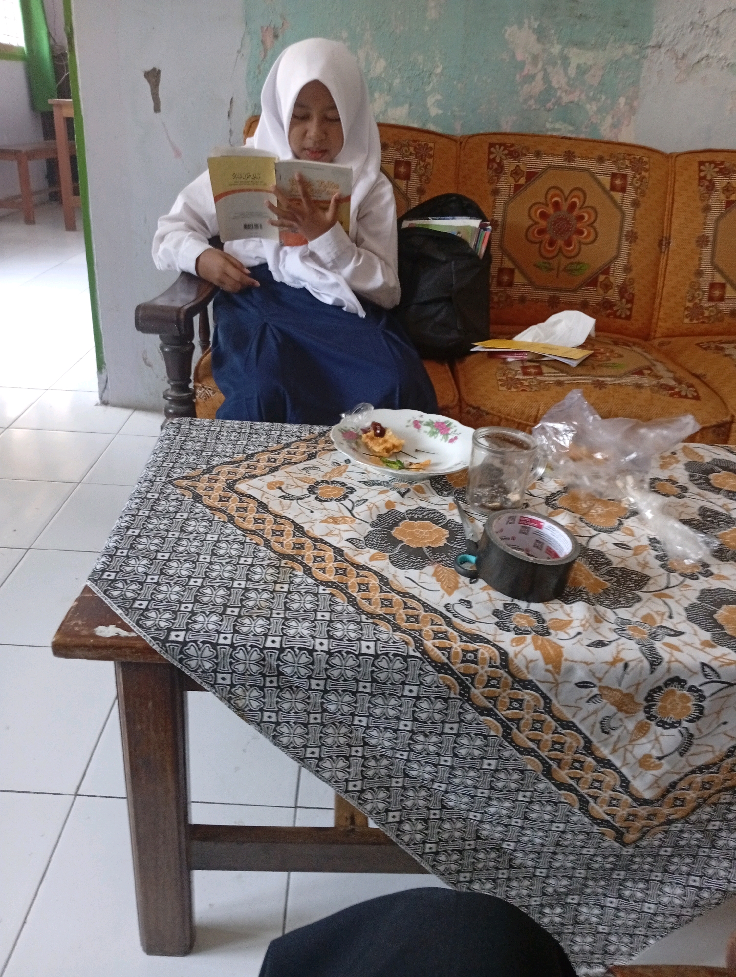 A young girl wearing a white hijab and a white shirt with a dark blue skirt is sitting on a wooden chair, reading a book. She is positioned in the foreground, on the left side of the image. Behind her, a section of a patterned, orange sofa is visible. A black backpack is placed on the sofa, to the right of the girl. In the mid-ground, a wooden table is covered with a batik patterned tablecloth. On the table, there is a small plate with food, a clear glass, a roll of black duct tape, and a pair of scissors. The background consists of a wall with peeling paint and a window with a green curtain. The floor is tiled. The overall scene appears to be an indoor residential setting, possibly in Gempeng, Indonesia, given the cultural context suggested by the attire and the batik pattern.