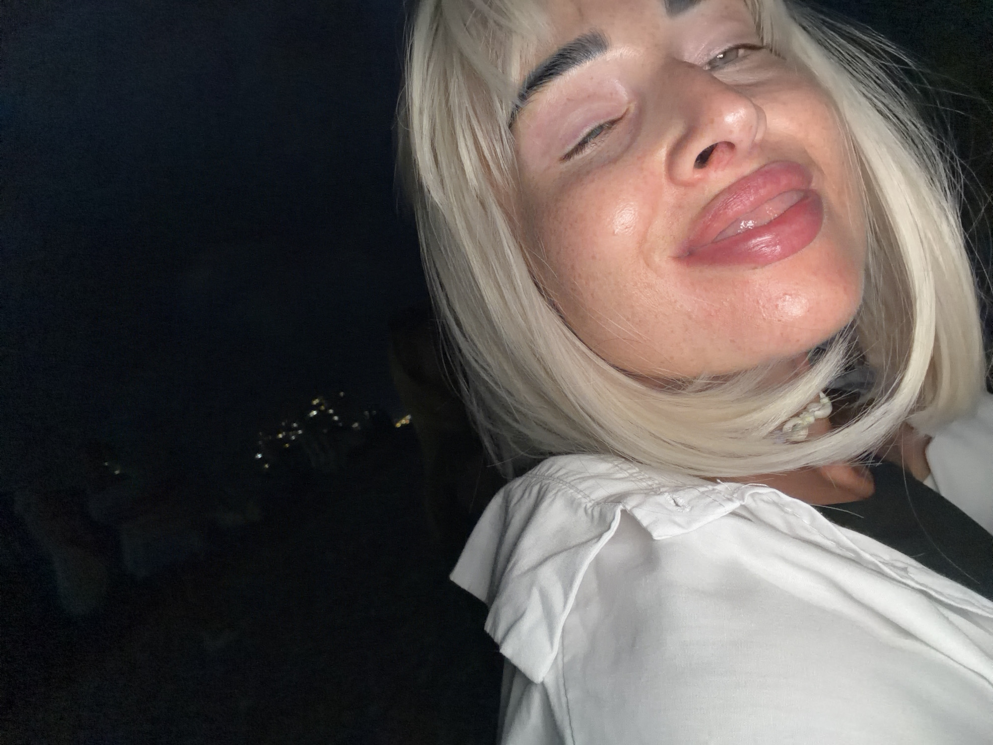 In the foreground of this low-light, high-angle image, a woman with platinum blonde hair and closed eyes is the primary subject. She is wearing a white collared shirt, partially buttoned, revealing a pearl necklace. Her lips are full and glossed, and her face shows signs of makeup. The background is predominantly dark, with faint, blurred lights visible in the distance, possibly representing city lights. The overall impression is a selfie taken at night, likely from a moving vehicle given the tilted perspective. The location is identified as Sovet-Kvadzhe, Russia, suggesting an urban nighttime environment.