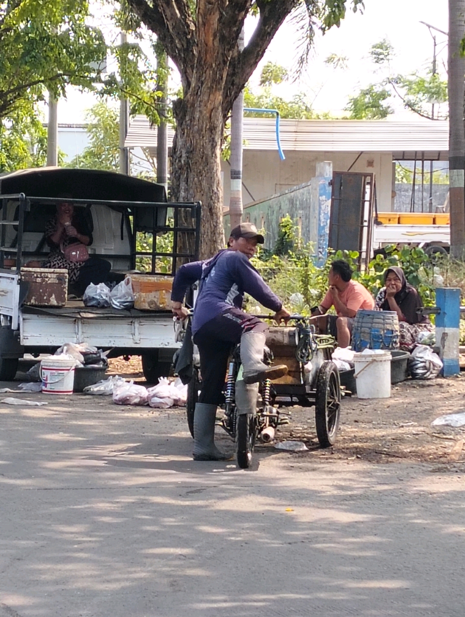 The image depicts a street scene in Probolinggo, Indonesia, likely a market area. In the foreground, a man wearing a blue long-sleeved shirt, dark pants, and grey boots is positioned on a three-wheeled motorcycle, appearing to be stopped or moving slowly. He is looking towards the left of the frame. Behind him, a white truck with a dark canopy is parked, with two women seated inside. One woman in the back is wearing a patterned dress and a pink bag, holding a white bottle. Another woman is seated to the right of the truck, wearing a headscarf and a patterned skirt.
Scattered on the ground around the truck and the motorcycle are various items, including white buckets, plastic bags filled with what appear to be goods, and a blue woven basket. A large tree with a textured trunk stands prominently on the right side of the frame, casting shadows on the asphalt road. In the background, there are buildings with white walls and a white-tiled roof, along with lush green foliage. The sky is bright and appears to be daytime. The overall impression is that of a bustling roadside market or a stop for vendors.