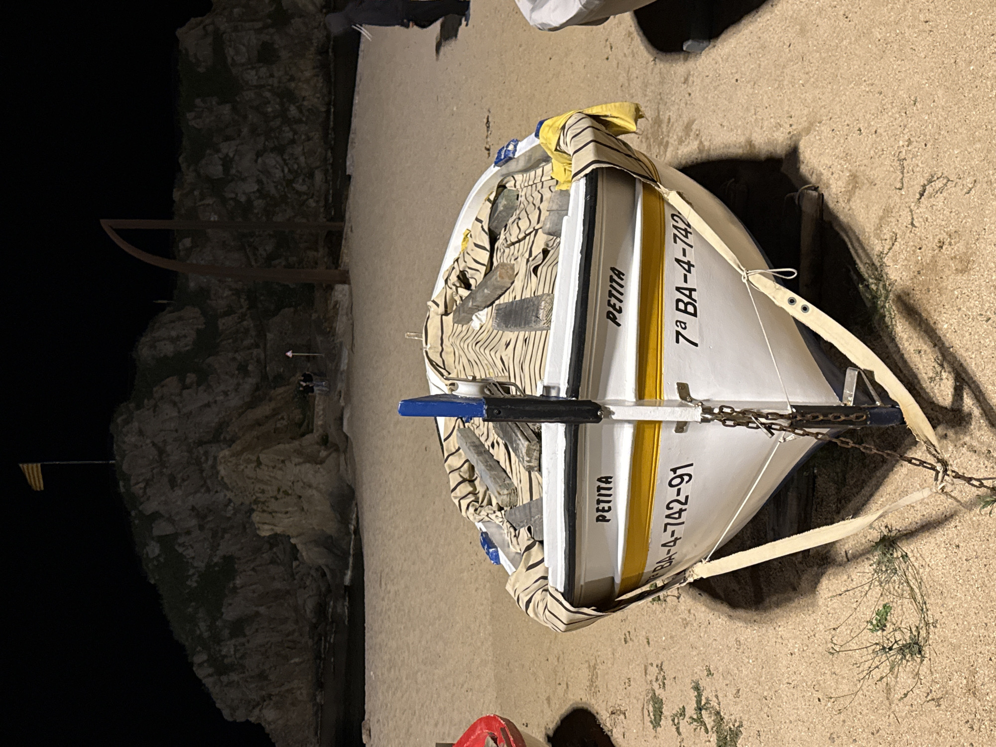 This image depicts a nighttime scene at the Blanes waterfront in Blanes, Spain. The foreground is dominated by a white fishing boat with yellow trim, resting on a sandy beach. The boat is partially covered with a striped tarp. On the side of the boat, the word "PETITA" is visible, along with registration numbers: "BA-4-742-91" and "7a BA-4-742". A chain and straps secure the boat to the sand. In the mid-ground, a body of water is visible, with waves gently lapping the shore. Beyond the water, a rocky cliff rises, topped by a Catalan flag. To the right of the cliff, a tall, dark, arched metal structure stands on the sandy area. A person wearing a dark hooded jacket is seen walking away from the camera on the right side of the frame. A second person, also wearing dark clothing, is visible in the distance near the base of the cliff on the left. The overall lighting suggests artificial illumination from nearby sources, casting shadows on the sand and highlighting the textures of the boat and the surrounding environment.