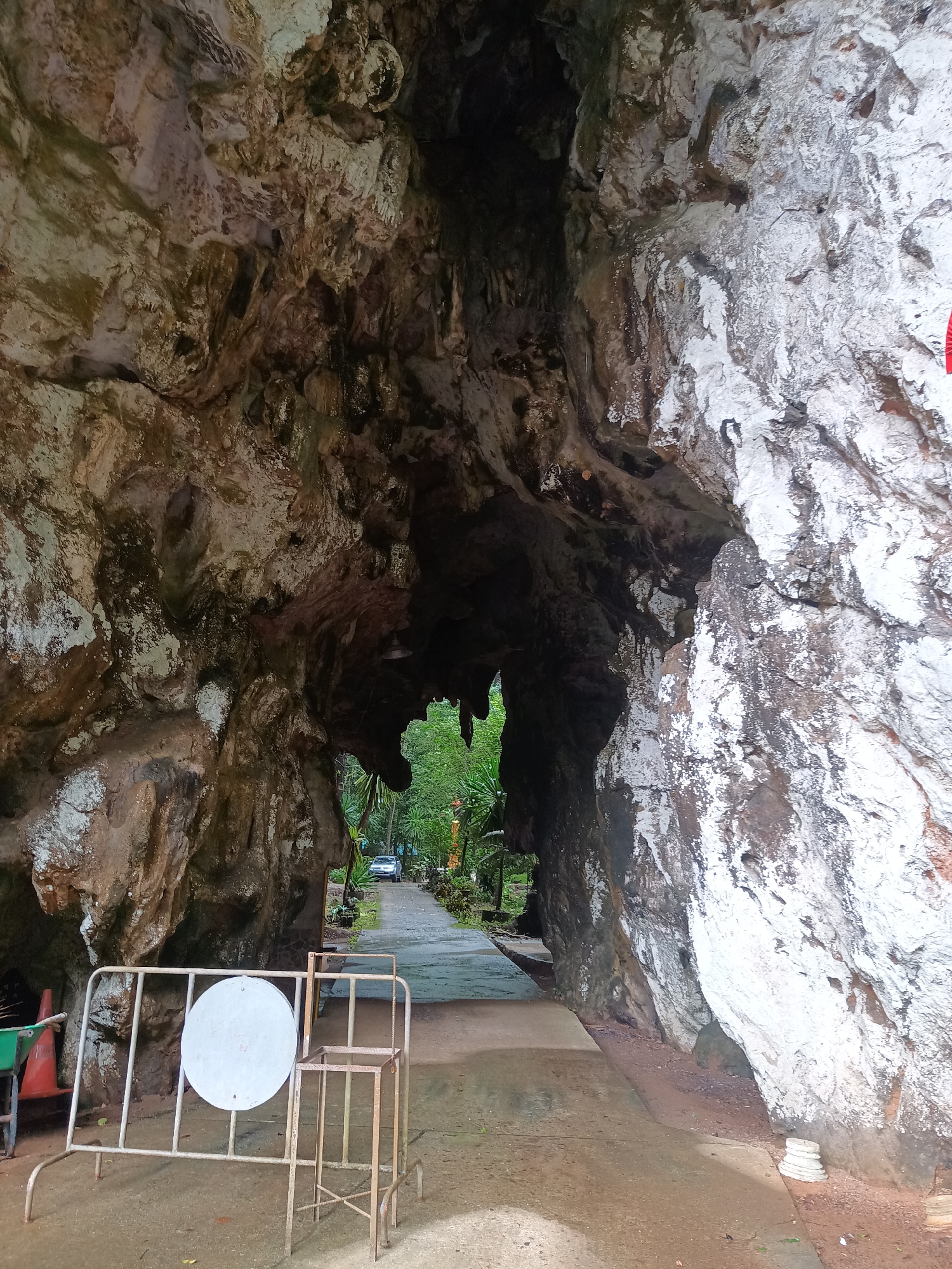 The image shows a cave entrance at Wat Tham Khao Plu temple in Ban Pak Ra, Thailand. The cave opening reveals a paved pathway leading to a distant vehicle and lush vegetation. A metal barrier with a white circular sign stands in the foreground. The cave walls are textured with a mix of light and dark stone.