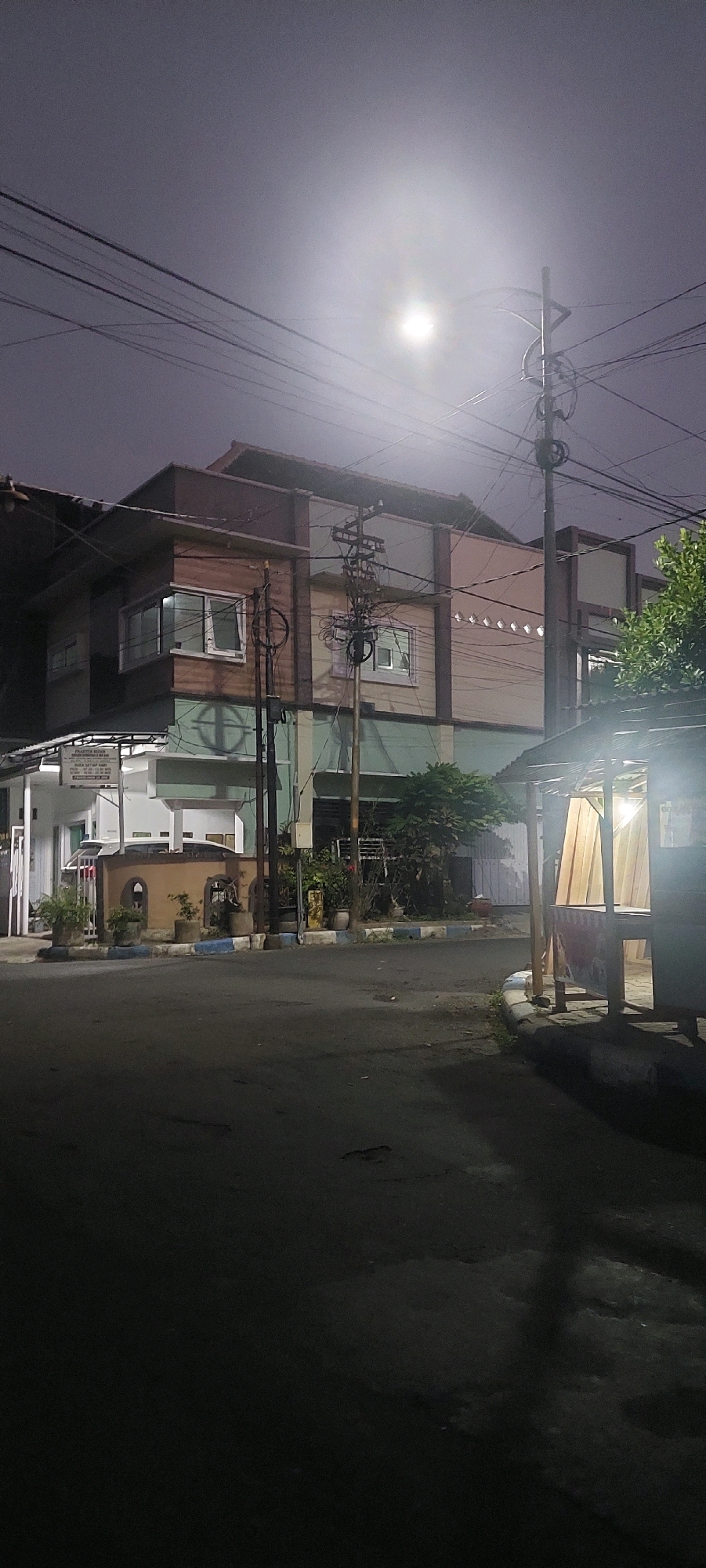 This is a nighttime photograph taken at a street corner in Lumajang, Indonesia. The scene is dimly lit by a bright, hazy light source in the sky, likely the moon obscured by clouds or fog, and by the lights emanating from a building and a roadside stall.
In the foreground, the asphalt street is visible, appearing dark and somewhat uneven. To the right, a small roadside stall or kiosk with a corrugated metal roof is illuminated from within, with light-colored curtains visible behind what appears to be a counter. The stall is constructed from wood and concrete.
To the left, a multi-story building dominates the mid-ground. The building has a modern design with wooden and colored paneling on its upper floors. Several windows are visible, some lit from within. A white sign with text is mounted on a lower section of the building. Electrical wires and poles are prominent throughout the scene, crisscrossing the sky and the building.
The environment appears residential or commercial. There are some potted plants and small trees around the building and along the roadside. The overall atmosphere is quiet and still, characteristic of a street at night. No people are visible in the frame.