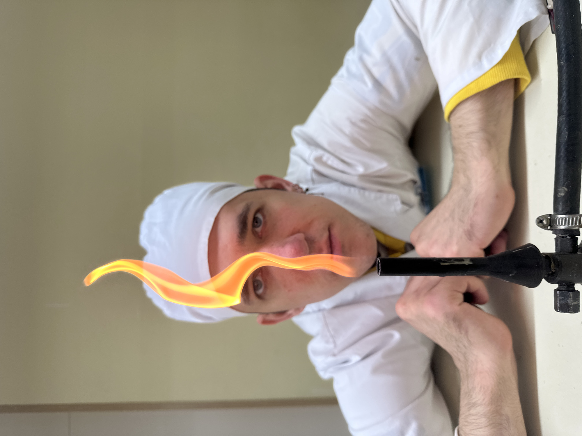 A man wearing a white chef's hat and white jacket with yellow trim is holding a lit gas burner. The background is a plain wall. I cannot determine the location in Saratov, Russia from this image.