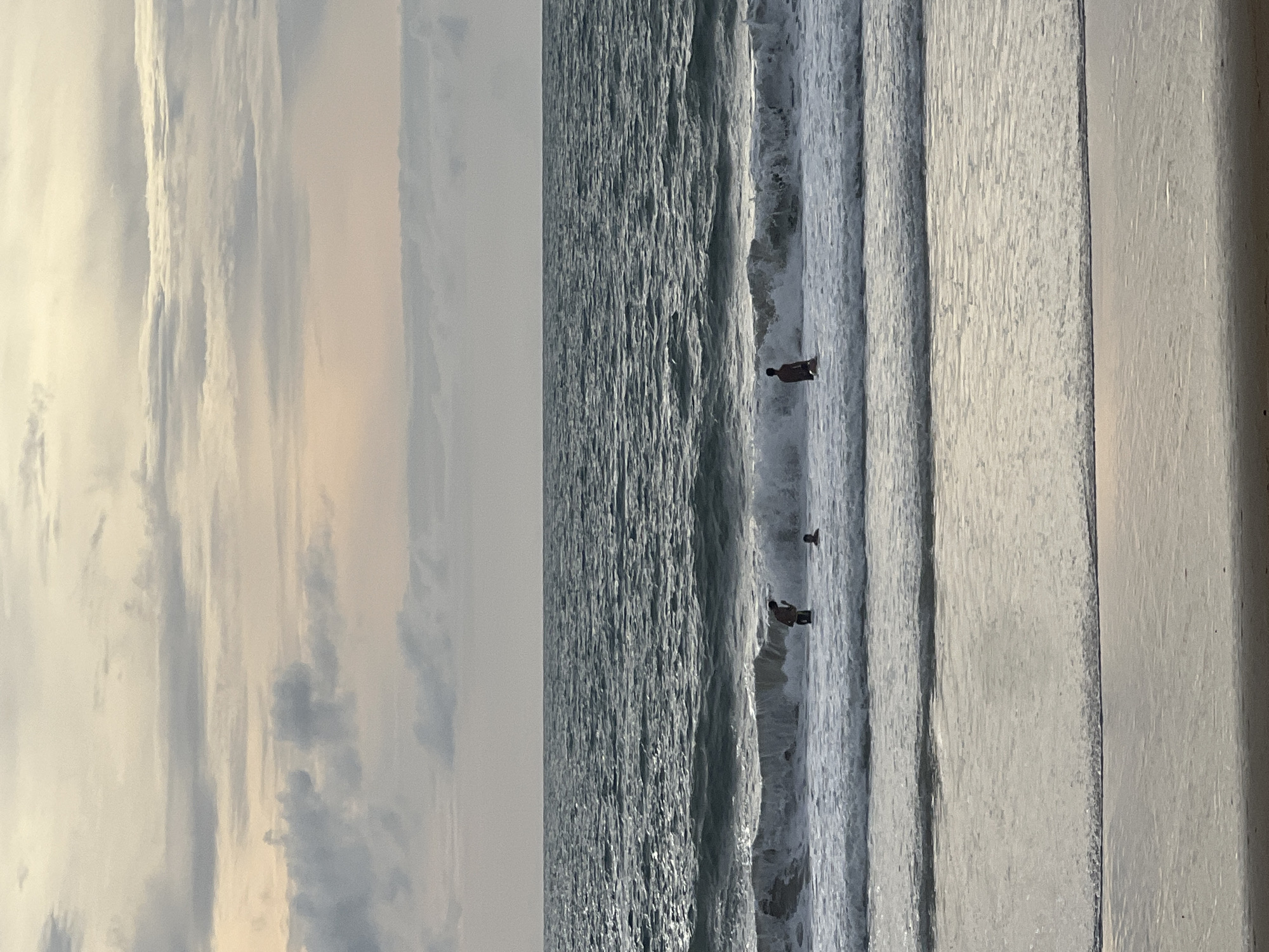 An ocean scene at dusk. Three individuals are visible in the water, with waves breaking around them. The sky is filled with clouds, tinged with pastel colors from the setting sun. The foreground shows the wet sand of a beach, reflecting the muted light.