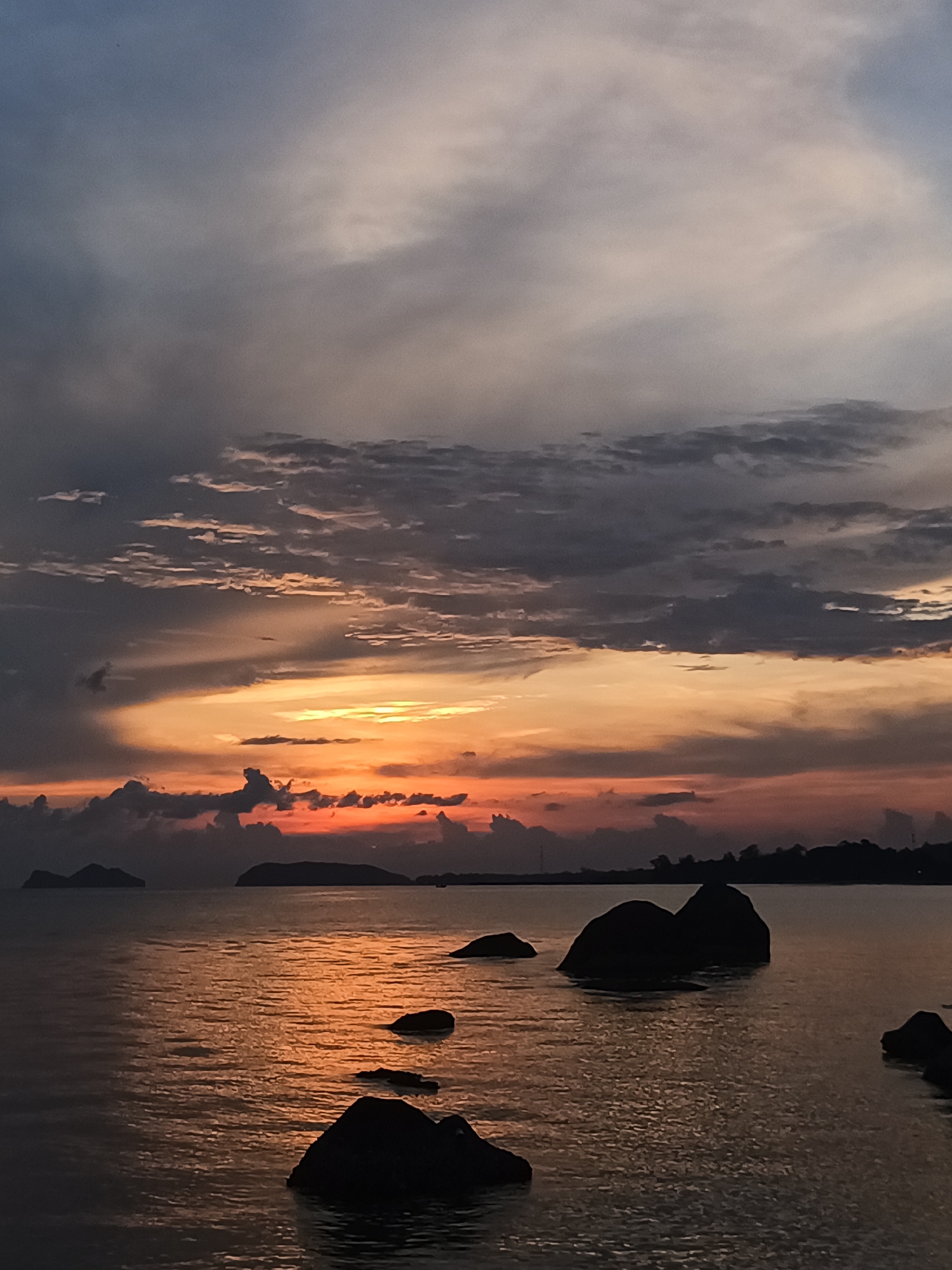 The image depicts a sunset over the water in Ban Haad Rin, Thailand. In the foreground, dark rocks are partially submerged in the water. The water reflects the orange and yellow hues of the sunset. In the distance, islands and the coastline are visible in silhouette. The sky is filled with clouds, ranging in color from dark gray to orange and yellow near the horizon.