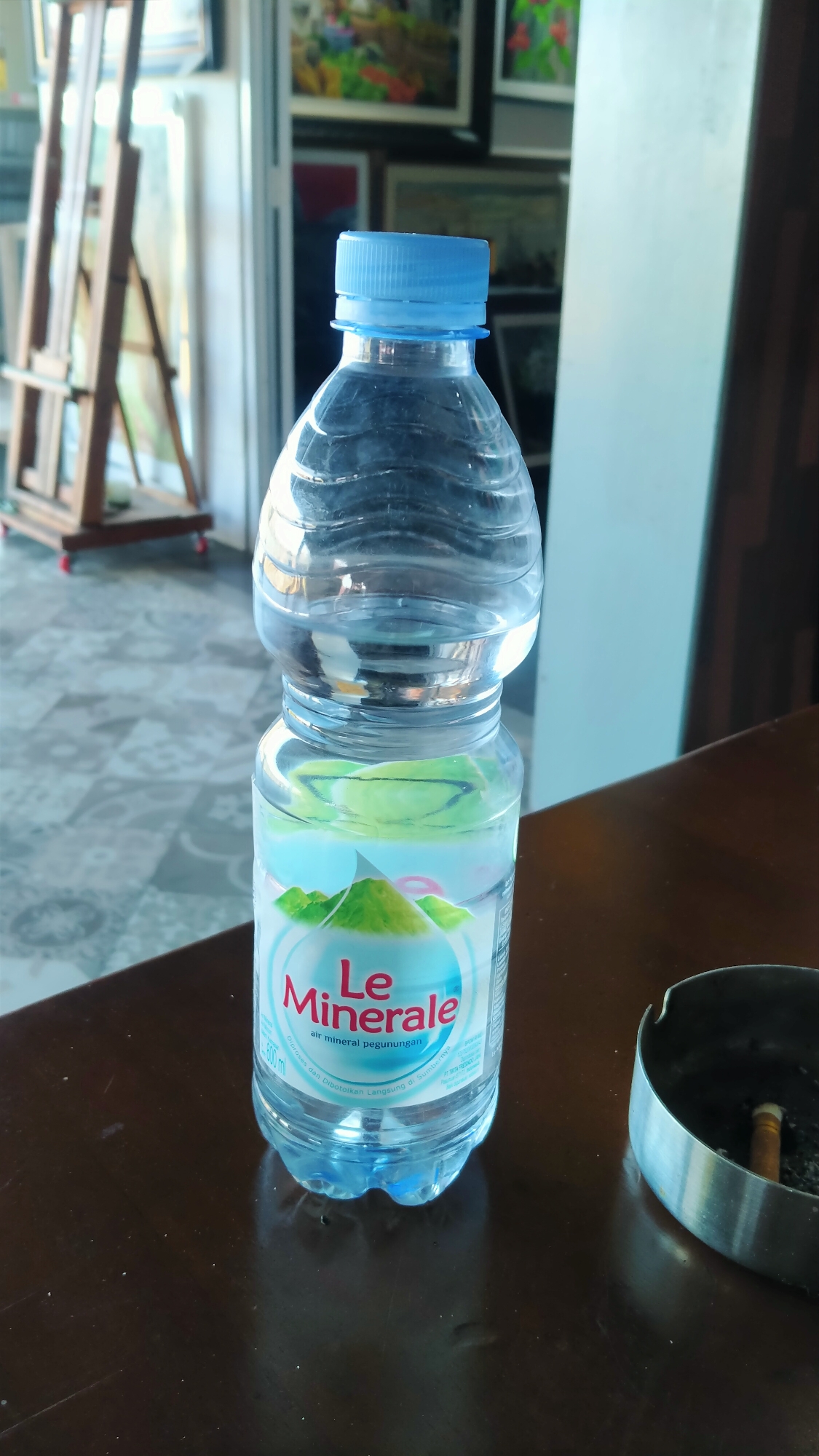 The image shows a clear plastic bottle of Le Minerale bottled water, with a blue cap, sitting on a dark brown wooden table. The bottle is partially filled with water. The label on the bottle features the Le Minerale logo, green mountains, and text in Indonesian. To the right of the bottle is a silver metal ashtray containing a lit cigarette. In the background, blurred elements suggest an indoor setting. To the left of the bottle, an easel with canvases is visible, and behind that, artwork is displayed on a wall. The floor in the background has a patterned tile design. The overall lighting suggests an indoor environment with natural light filtering in. The scene depicts a still life with a drink and smoking paraphernalia. It is impossible to determine the location in the city of Nyamat, Indonesia, from the provided image.