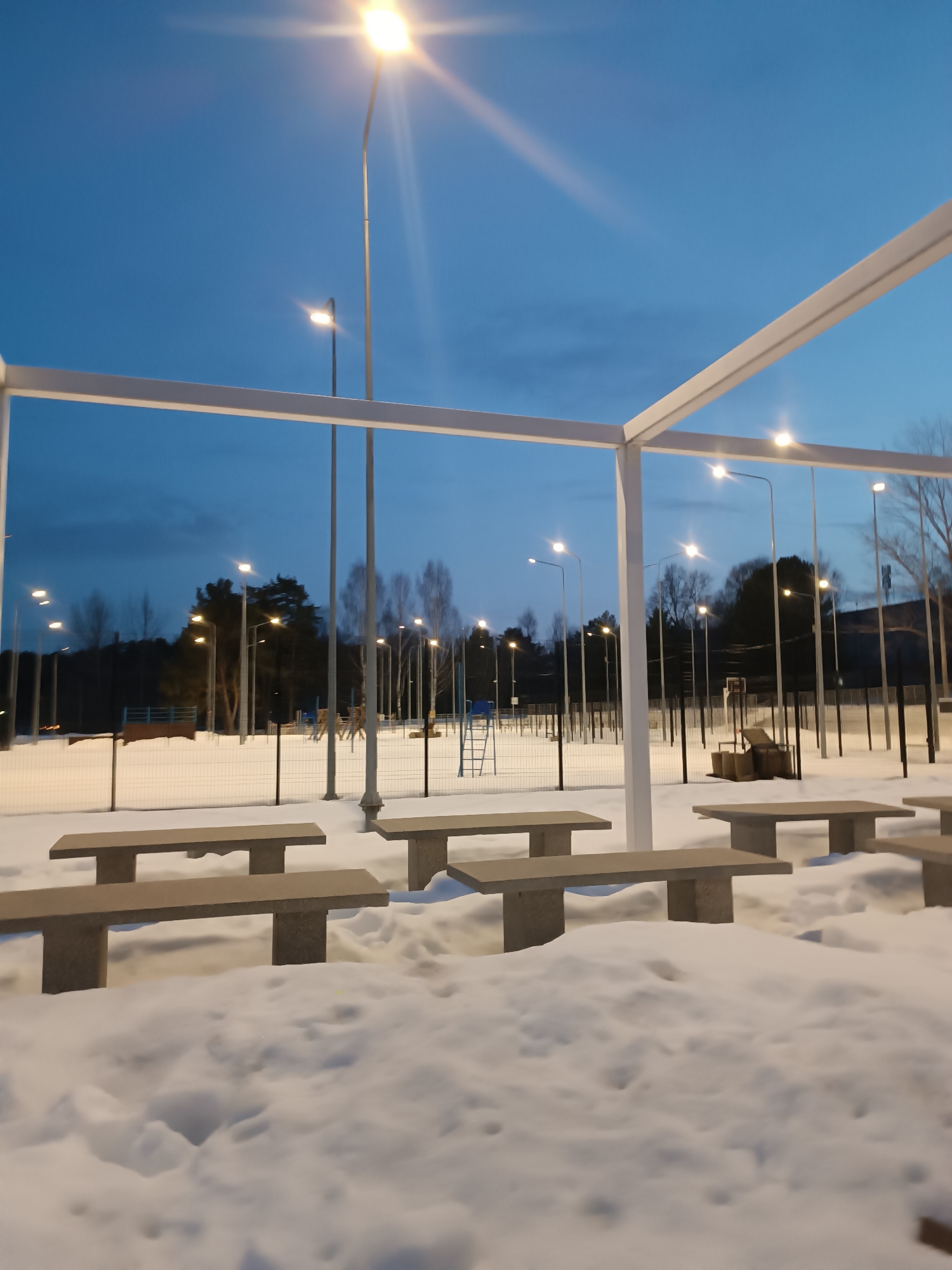 The image depicts an outdoor recreational area or park in the city of Unknown, Unknown, at twilight or early night during winter. The scene is illuminated by numerous artificial light sources against a deep blue sky. In the immediate foreground, a white, open-frame pergola structure partially frames the view. Beneath the pergola, the ground is covered in undisturbed snow, burying the bases of several light gray, rectangular concrete picnic tables and benches.
Beyond the tables, a snow-covered, fenced-off area is visible. The fence consists of black vertical posts supporting a white mesh barrier. Within this enclosed area, several tall streetlights with bright yellow-orange lamps illuminate the snow-covered ground and various structures. To the left, a blue-colored playground slide structure is partially visible. To the right, a basketball hoop with a brown wooden backboard and a collection of dark cylindrical objects (possibly bins or weights) are present.
The background consists of a dense line of trees, both coniferous and bare deciduous, appearing dark against the sky. More streetlights are visible among and behind these trees. No individuals are present in the depicted scene, and no discernible actions or interactions are occurring. The environment is characterized by snow cover and artificial illumination under a clear, dark blue sky.