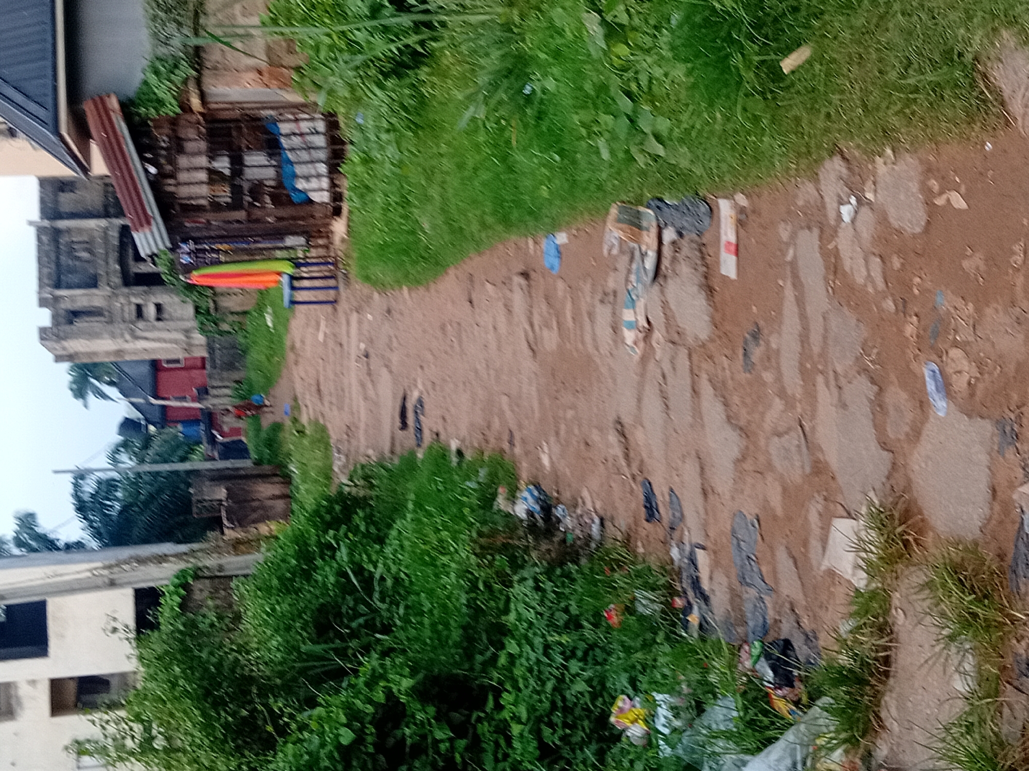 The image depicts a dirt path in Ifite-Awka, Nigeria, flanked by overgrown green vegetation on the left and a grassy area on the right. The path is uneven and appears to be constructed from compacted earth and stones, with scattered debris and litter along its edges and on its surface. On the left side of the path, there is dense green foliage, and further back, a partially visible building with a white facade. On the right side, there is a small, makeshift stall with a corrugated metal roof, its front covered by a metal grate. Several colorful fabrics, possibly garments, are hanging from a stand in front of the stall. In the background, there are more buildings, some appearing partially constructed or in disrepair, and utility poles with wires. The sky is overcast. In the mid-ground, two individuals are walking away from the camera down the path, one carrying a blue bag. The overall scene suggests a somewhat neglected urban or semi-urban environment.