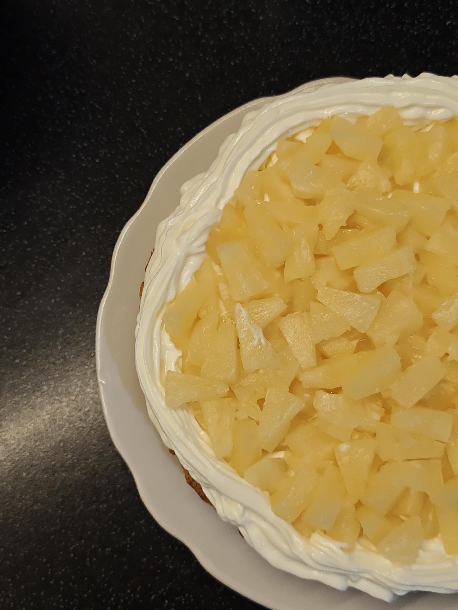 A high-angle shot of a cake on a white plate resting on a dark, textured surface. The cake is decorated with concentric rings of white whipped cream and is topped with uniformly chopped pineapple. The environment is minimalist, focusing attention on the cake. No location or other subjects/objects are apparent.