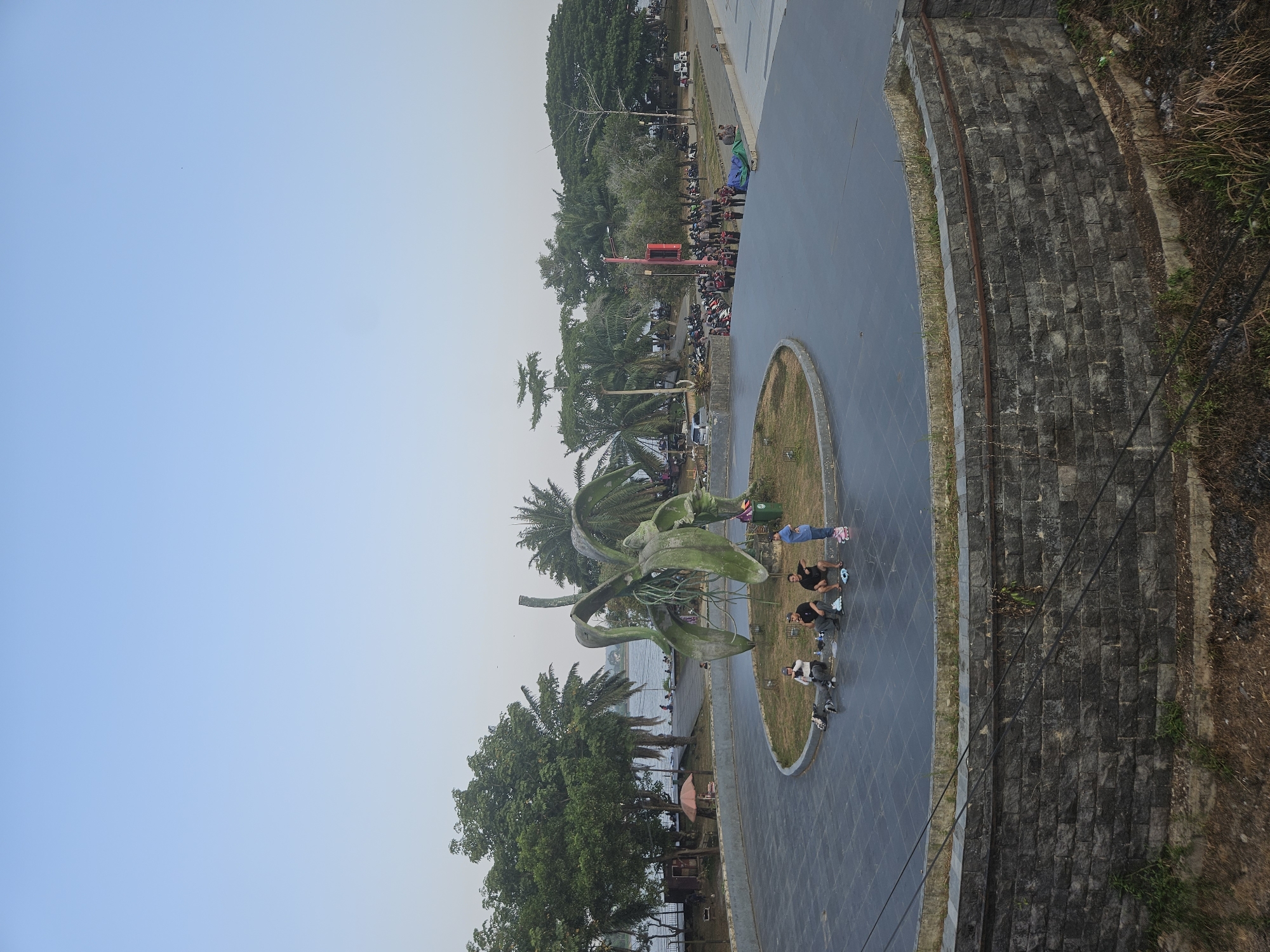 The image depicts a public park or plaza in Teluk Dalam, Indonesia, with a large, abstract metal sculpture resembling a plant or flower in the foreground. The sculpture is green and weathered, with visible roots extending from its base. Several people are seated and standing around the base of the sculpture, some on the paved area and others on the grassy median. One person is wearing roller skates, and another is wearing roller blades.
Behind the sculpture, a paved walkway curves around a body of water, likely a lake or river, with trees and palm trees lining the bank. In the middle distance, there is a parking area with numerous motorcycles and a few cars. A group of people, some wearing uniforms, are gathered near a red pole with a sign. Further in the background, more trees and buildings are visible under a clear, pale blue sky. The foreground features a textured stone wall. The overall scene suggests a leisurely outdoor gathering spot.