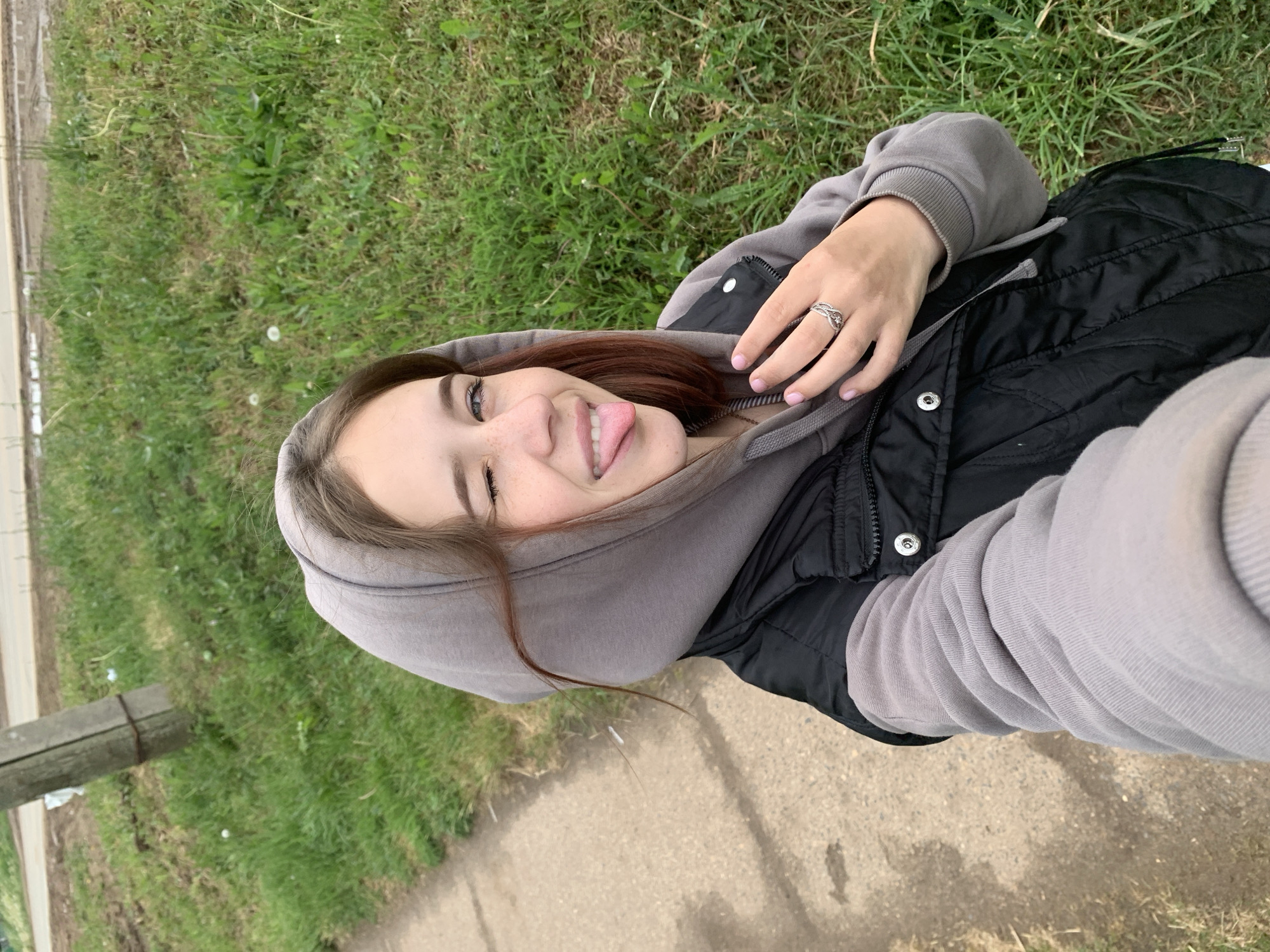 A young woman is taking a selfie outdoors. She is wearing a grey hoodie and a black vest. She is sticking her tongue out and smiling. The background consists of a grassy area and a sidewalk. The location is near Фоки, Russia.