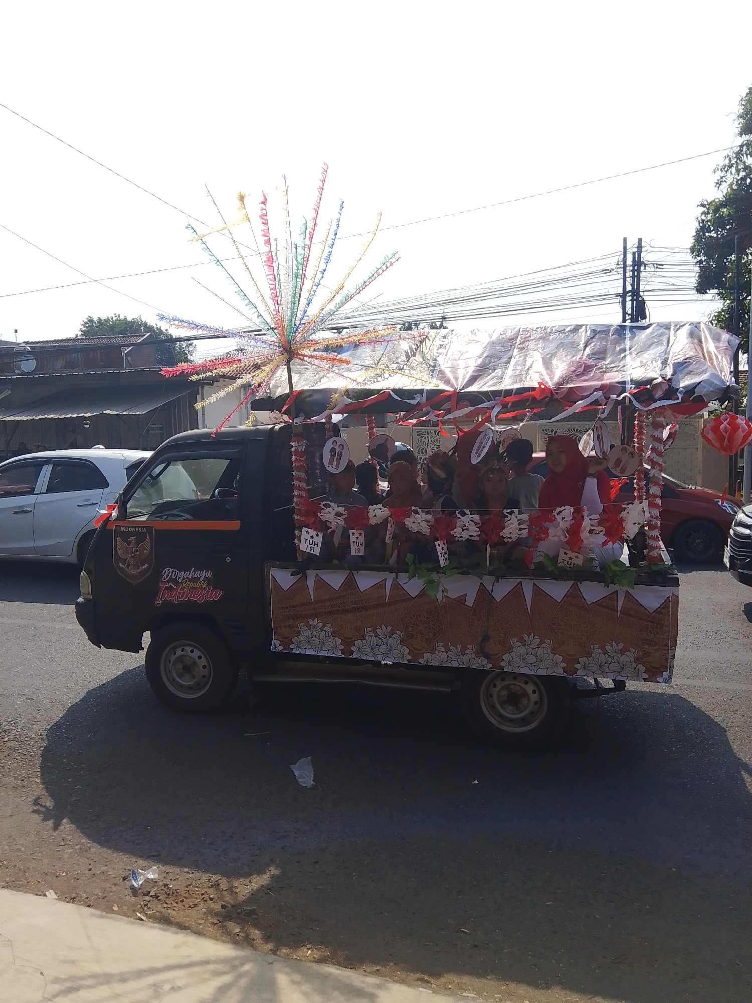 In Kalinyamatan, Indonesia, a black pickup truck, decorated with Indonesian flags and patriotic imagery, participates in a parade. Children and adults are seated in the back of the truck, which is adorned with a canopy made of white and red fabric, streamers, and paper decorations. The truck itself features a large emblem on its side with the text "INDONESIA" and "Dirgahayu Republik Indonesia." The vehicle is driving on an asphalt road, with a white car and a red car visible in the background, as well as buildings and trees lining the street. The sky is clear and bright, suggesting a sunny day. The scene conveys a celebratory atmosphere, likely related to Indonesian Independence Day.