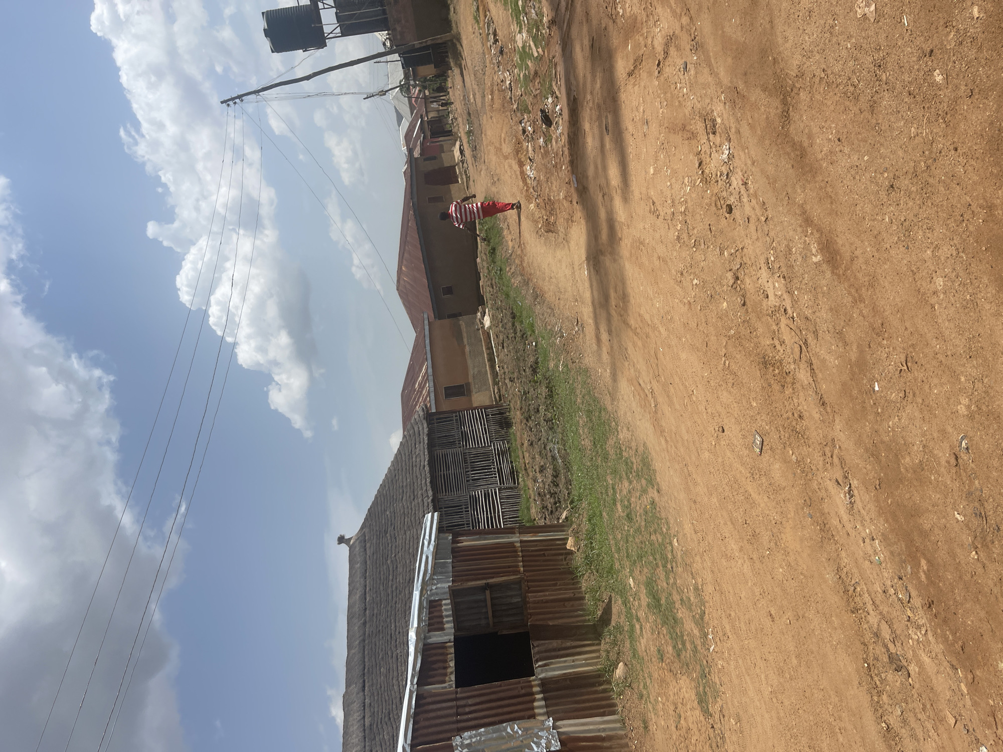 The image depicts a rustic scene in Rafin Par, Nigeria. The foreground is dominated by a dirt road, which appears dry and dusty with patches of sparse green grass along its edges. To the left of the road, several structures are visible. One is a building with a corrugated metal roof and walls, featuring an open doorway. Adjacent to it is another building with a thatched roof and wooden slat walls. Further down the road, more buildings with red metal roofs and light-colored walls can be seen.
A person wearing a red and white striped shirt and red pants is walking along the dirt road, facing away from the camera. In the background, a water tank is mounted on a tall structure, supported by a pole with several electrical wires stretching across the frame. The sky is partly cloudy, with large white cumulus clouds against a blue sky, suggesting it is daytime. The overall impression is one of a rural or semi-urban environment in Nigeria, with a simple, functional style of construction and a modest way of life. There is no visible text in the image.