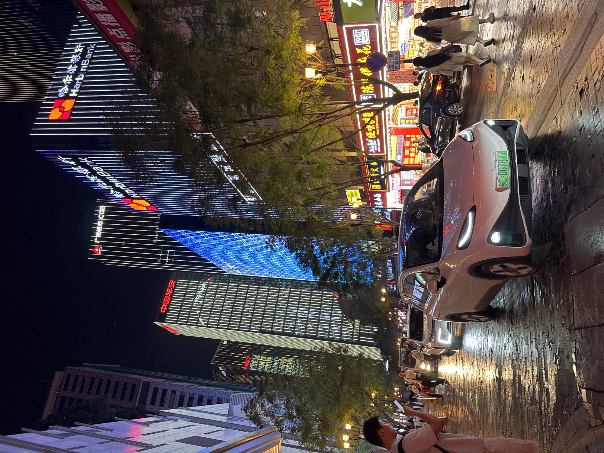 The image depicts a nighttime street scene in the Yuzhong District of China, likely a commercial area due to the presence of illuminated signs and tall buildings. Several pedestrians are visible, and cars are parked along the street. One prominent white car is in the foreground. Buildings with illuminated signs, including "Harbin Bank," are visible in the background. A person is taking a photo with their phone. The road surface appears wet.