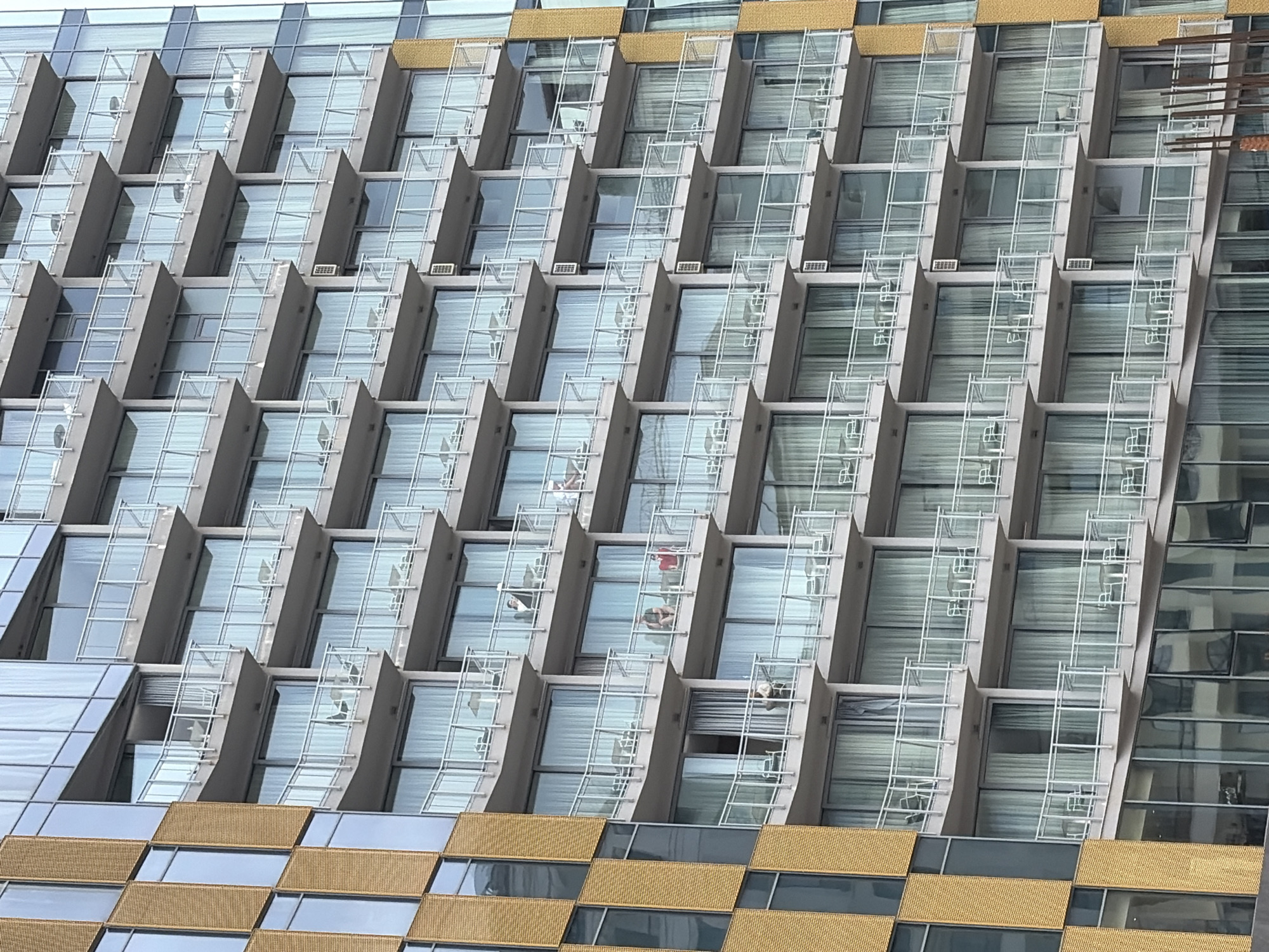 The image shows a close-up of a modern building facade, possibly a hotel or residential complex, located in Rafailovići, Montenegro. The building features a repeating pattern of cantilevered balconies with large glass windows. The balconies are divided by grey vertical structures. Some windows have white blinds drawn down. The facade has alternating gold and mirrored glass panels on the lower portion of the building. The lighting suggests an overcast or partly cloudy day. There are people visible on some of the balconies.
