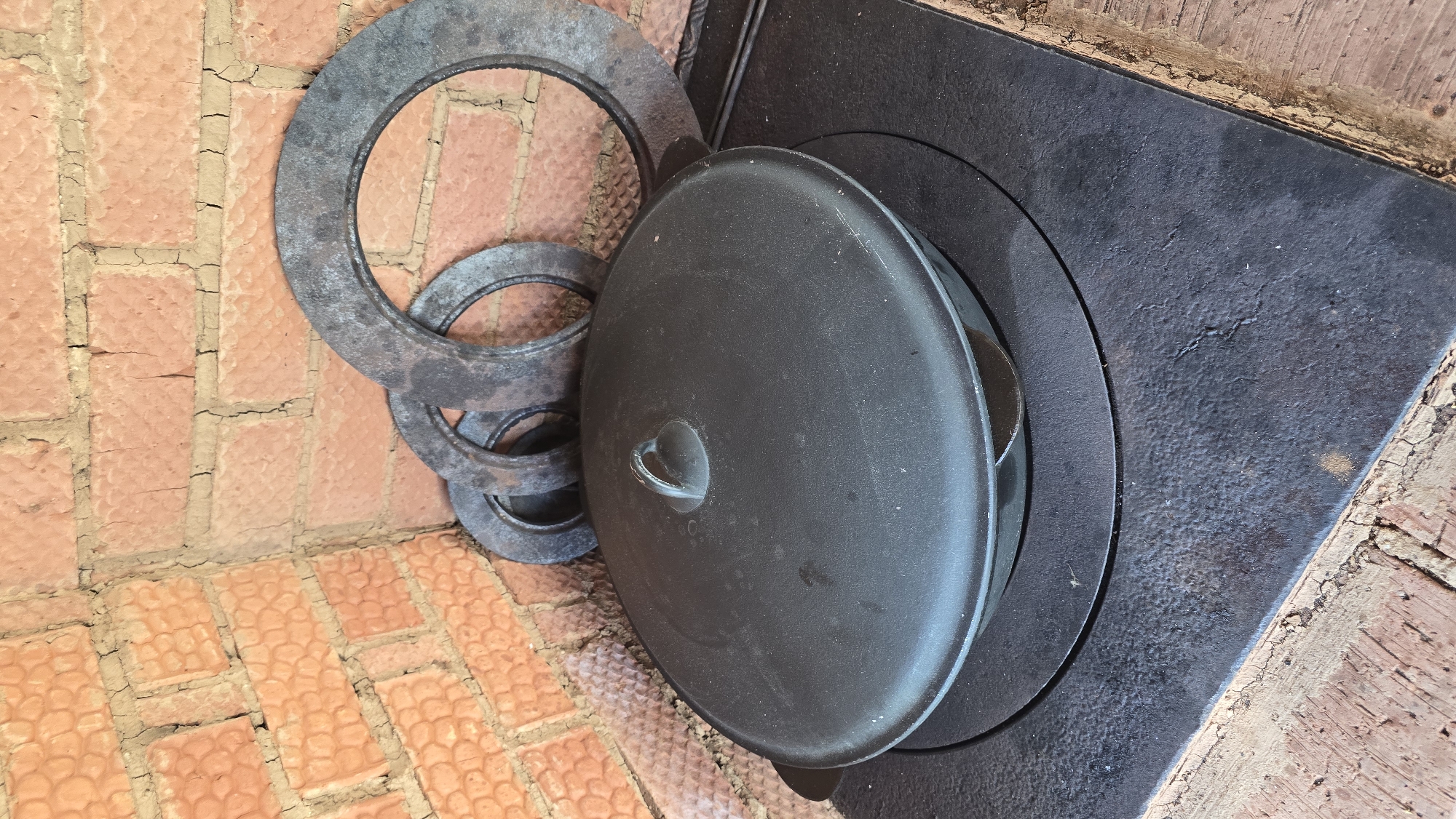 The image depicts a cast-iron cooking setup situated in Забегалово, Russia. A large, dark grey cast-iron pot with a fitted lid and a central loop handle rests on a black, rectangular cast-iron stovetop. The stovetop features a circular indentation where the pot is seated, suggesting it's part of a traditional cooker or wood-burning stove. Behind the stovetop, three concentric, dark metallic rings of varying diameters lean against a rustic brick wall. The wall consists of reddish-orange bricks with textured surfaces and irregular, cracked mortar joints, forming an interior corner. No individuals or active processes are visible in the scene.