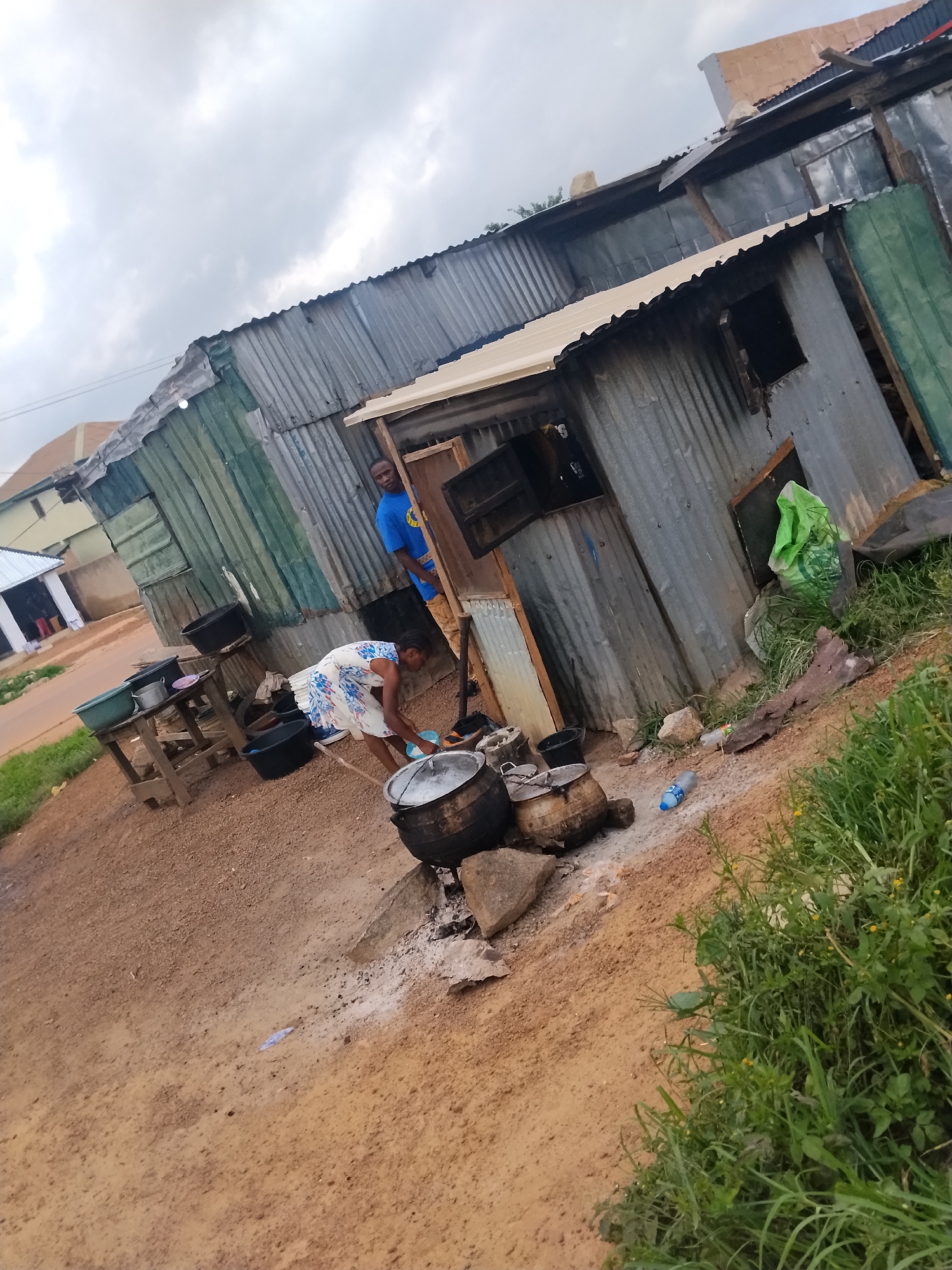In Durumi, Nigeria, a woman in a floral dress is bent over, tending to a large pot on a makeshift stove outside a corrugated metal structure. She appears to be washing or preparing something in the pot. Nearby, a man in a blue t-shirt stands and observes the activity. The environment consists of a dirt ground, some grass, and other assorted objects including buckets, smaller pots, a wooden table, and a plastic bottle. The structures are rudimentary, indicating a less developed area. The sky is overcast.