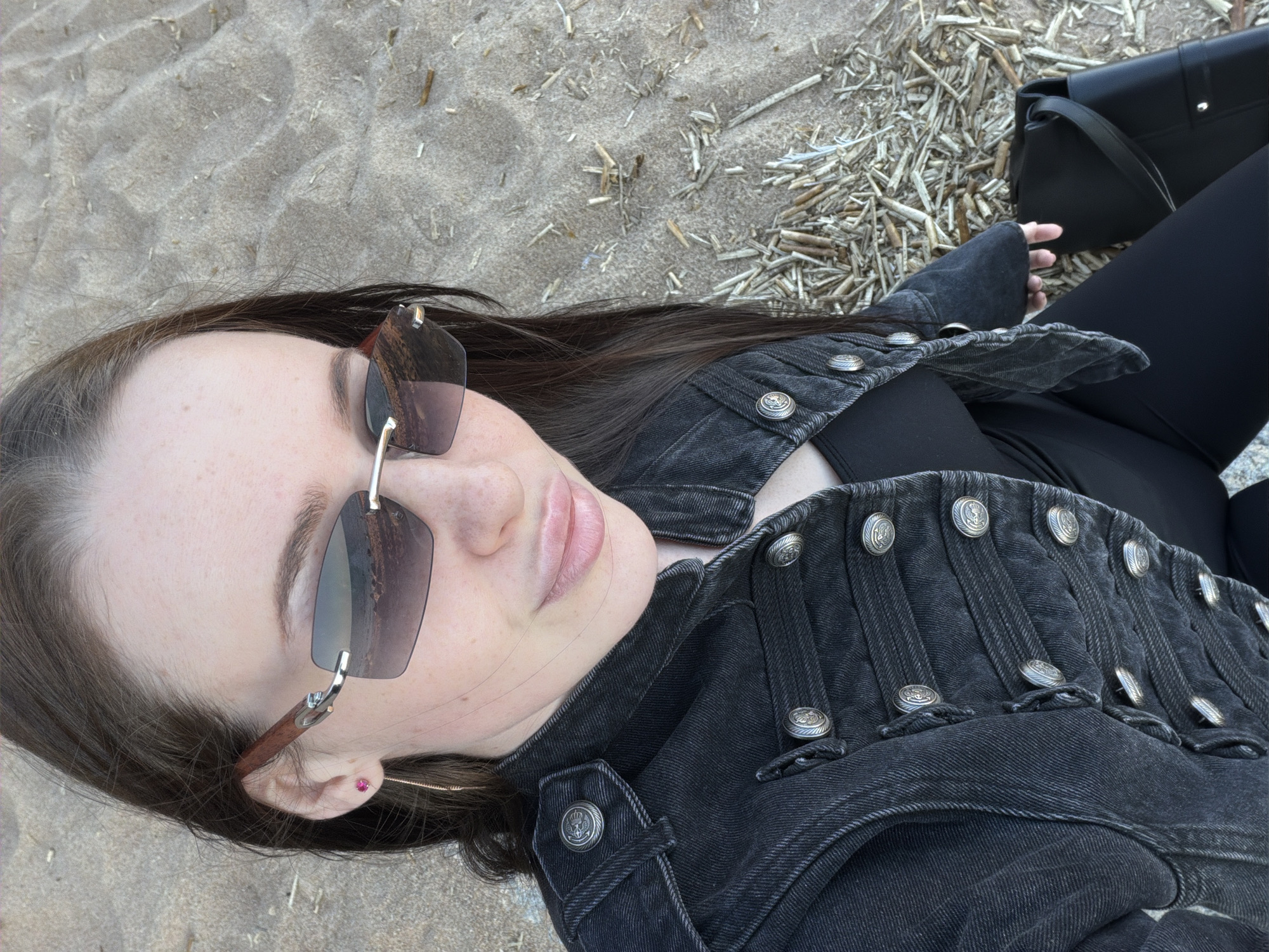 A female subject is depicted in a medium-shot, from a slightly elevated perspective, seated on a sandy surface identified as a beach in Repino, Russia. The subject has long, dark brown hair and fair skin with visible freckles on the forehead and cheeks. She is wearing dark-tinted, rimless sunglasses with reddish-brown, possibly wooden, temples that reflect a body of water and a distant horizon. On her visible left ear, she wears a small pink stud earring and a thin, golden-toned hoop earring. She is dressed in a dark grey or black denim jacket featuring a military-style design with multiple horizontal straps and numerous silver-toned, embossed buttons across the chest, shoulders, and cuffs. A black top is visible underneath the jacket, and she is wearing black leggings or tight-fitting trousers. A black, structured shoulder bag with a silver-toned stud is positioned beside her right leg. The ground is composed of light-colored sand with discernible ripples and is littered with scattered dried, light-colored reeds and plant debris, particularly on the right side of the frame. The subject is looking directly towards the camera with a subtle smile.