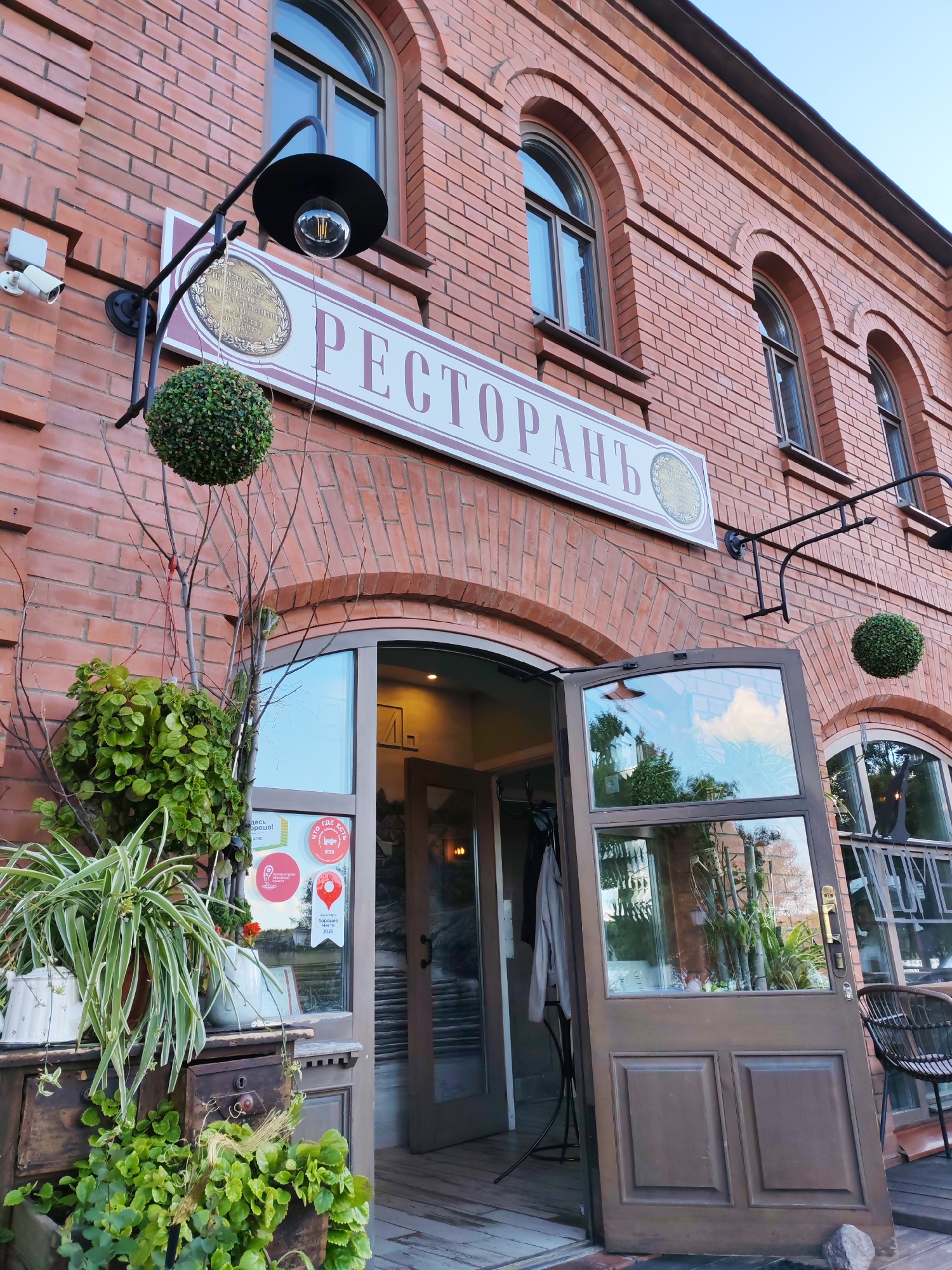 This media file shows the exterior of a restaurant in Plyos, Russia. The building is made of red brick and has arched windows. Above the entrance, a sign reads "РЕСТОРАНЬ" (Restaurant). The entrance consists of double doors, one of which is open, revealing a glimpse of the interior.
The scene is decorated with greenery, including potted plants on a rustic wooden dresser next to the entrance and a spherical topiary hanging from an ornate lamp fixture. A security camera is mounted on the brick wall to the left of the entrance. The lighting suggests it is either early morning or late afternoon, with natural light illuminating the scene and a warm glow emanating from inside the restaurant. The sky is clear and blue, indicating fair weather.
On the window next to the open door, there are several stickers, including a red location pin icon, a circular emblem with text, and other small circular stickers. One visible sticker has the text "ЧТО ГДЕ ЕСТЬ" (What Where Is) and "Хорошее место 2026" (Good Place 2026).
The overall impression is one of a welcoming and established restaurant with a charming, slightly rustic ambiance. There are no people visible in the frame.