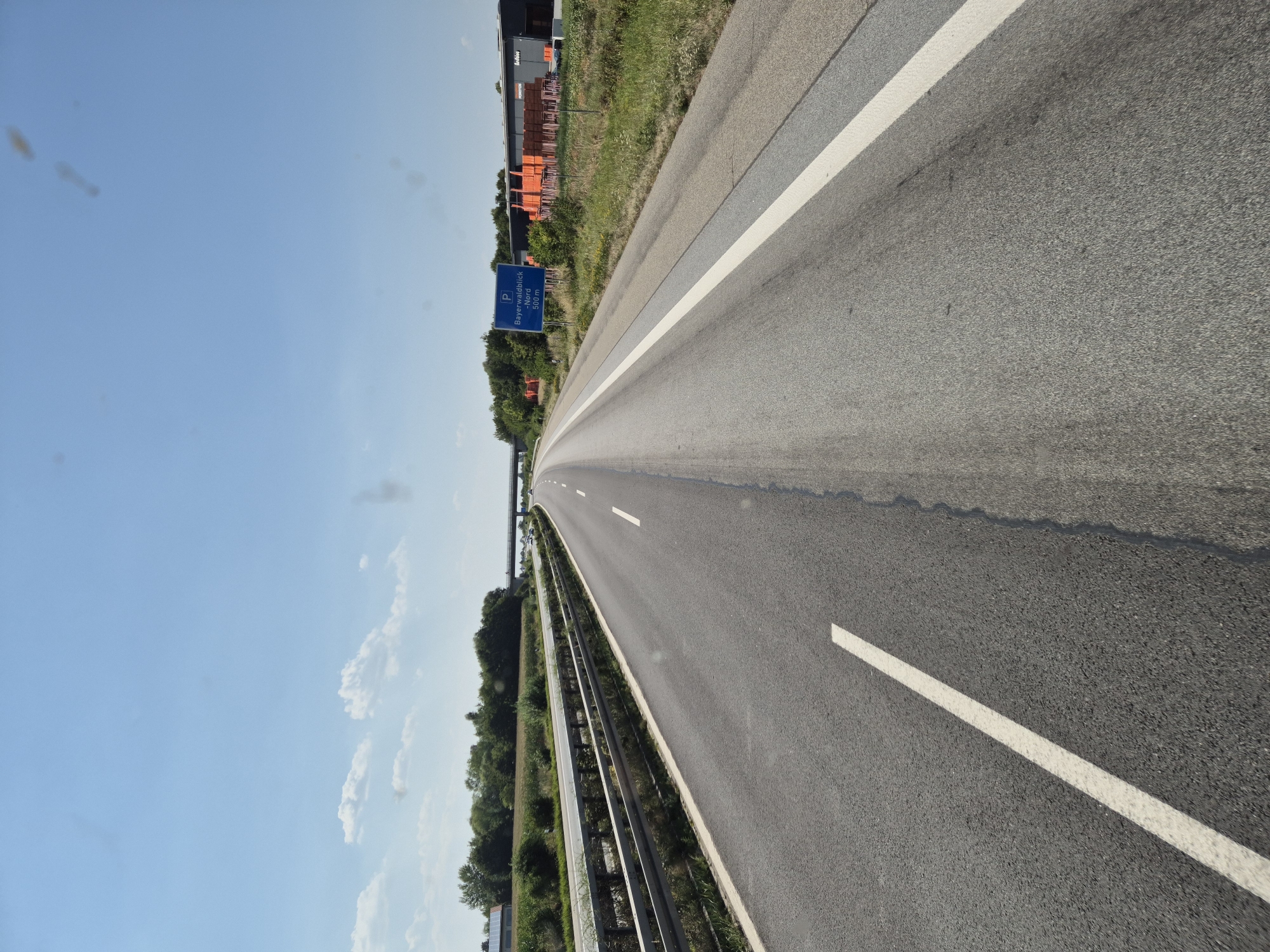 The image shows a highway scene in Germany on a clear, sunny day. The perspective is from a vehicle moving forward on a multi-lane highway. The road has asphalt surfacing with distinct white lane markings. To the left of the road is a metal guardrail separating the highway from a grassy area and a field with some trees in the distance. Further in the distance on the left, there are buildings, possibly agricultural or industrial. On the right side of the highway, there is a grassy embankment with some vegetation. A blue highway sign is visible on the right, indicating "P Bayerwaldblick -Nord 500 m". Beyond the sign, there is a building with stacked orange materials in front, suggesting a commercial or industrial area. The sky is blue with a few scattered clouds. The overall scene depicts a typical highway environment. Based on the sign, the location is near Plattling, Germany, specifically related to the "Bayerwaldblick - Nord" rest stop or viewpoint, which is 500 meters ahead. There are no people or animals visible in the image.