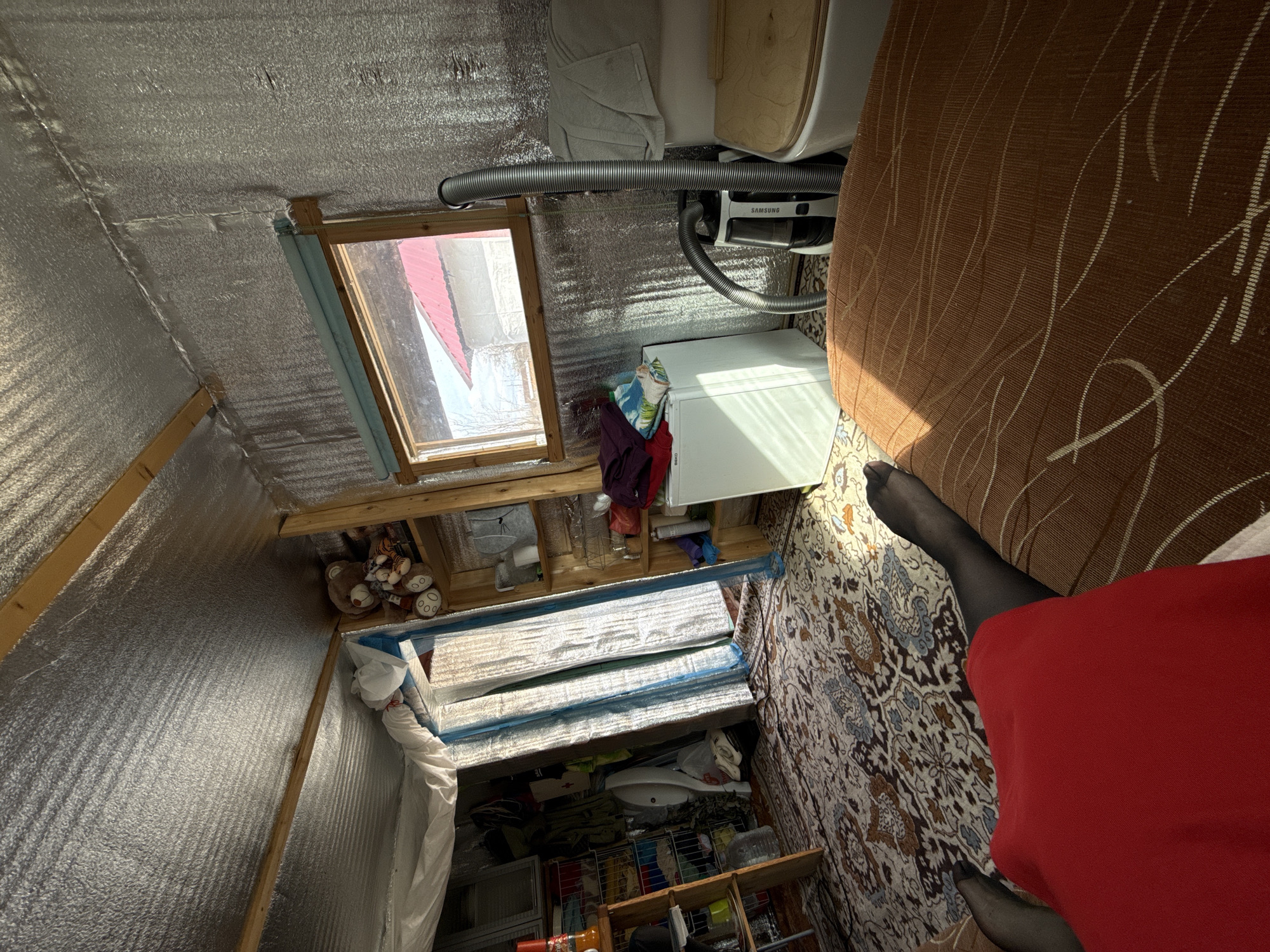 A distorted, high-angle view inside what appears to be an insulated attic in Chaikovsky, Russia. The ceiling and walls are covered with a reflective, foil-like insulation material. A skylight is present. Beneath the skylight are shelves containing toys and various objects. A Samsung vacuum cleaner is attached to a nearby wall. A section of brown upholstered furniture is visible in the upper right. Part of a person wearing black tights and a red dress is in the foreground. There is a rug on the floor. The scene conveys a cluttered and unconventional storage space within a residential dwelling.