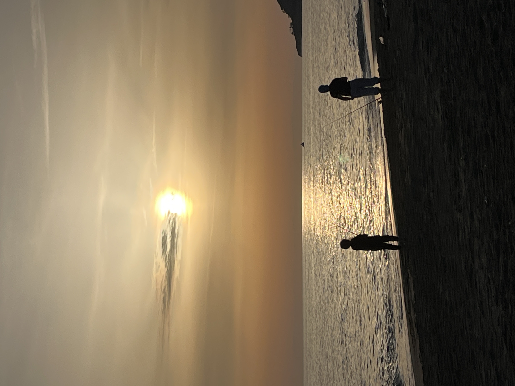 Two people are fishing in the sea at sunset. The sun is setting on the horizon, casting a warm glow over the water. The silhouettes of the people are visible against the bright background. The location is Alanya, Turkey.