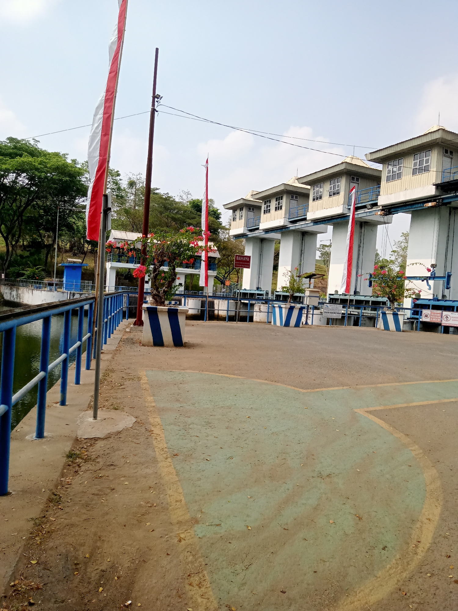 This is an outdoor, eye-level, full shot of a dam structure with a body of water on the left side and a pathway in the foreground. The dam has a series of white, rectangular buildings with windows, elevated on large white pillars. In front of the dam, there are red and white flags planted along a pathway. The pathway is paved with light brown material, with green and yellow markings. There are blue railings along the left side of the pathway, separating it from the water. Trees with green foliage are visible in the background, behind the dam. The sky is overcast with some white clouds. The location is identified as the Tiudan Dam in Tiudan, Indonesia.