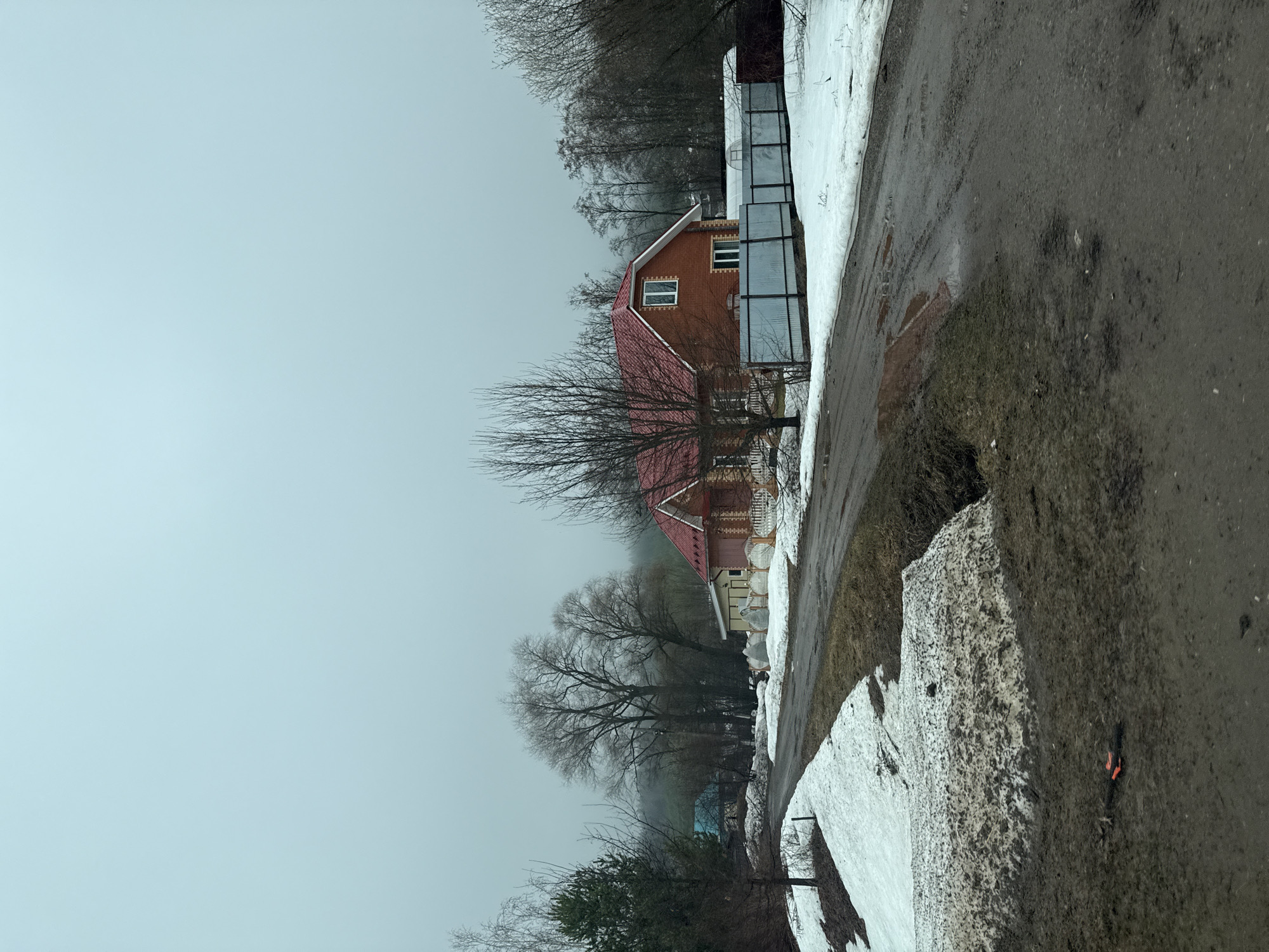The image depicts a scene in Гремяча, Russia on an overcast day with visible snow accumulation. A brick house with a red roof stands prominently, partially obscured by bare trees. Adjacent to the house is a metal fence or structure. A road or path borders the scene, showing remnants of melted snow and muddy conditions. The sky is uniformly gray, suggesting a bleak or wintry atmosphere. In the foreground, piles of melting snow are visible along the roadside. The overall impression is one of a rural or semi-rural setting in late winter or early spring.
