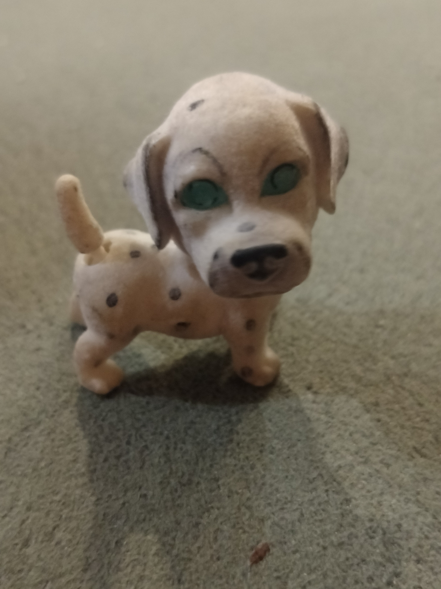A close-up, high-angle, soft-focus shot shows a small, felt-like toy dog. The dog is primarily cream-colored with black spots and has bright green eyes. It is standing on a textured, grey surface that appears to be carpet. The lighting is dim, casting a shadow of the dog to its right. No location in Novosibirsk, Russia is discernible from the image.