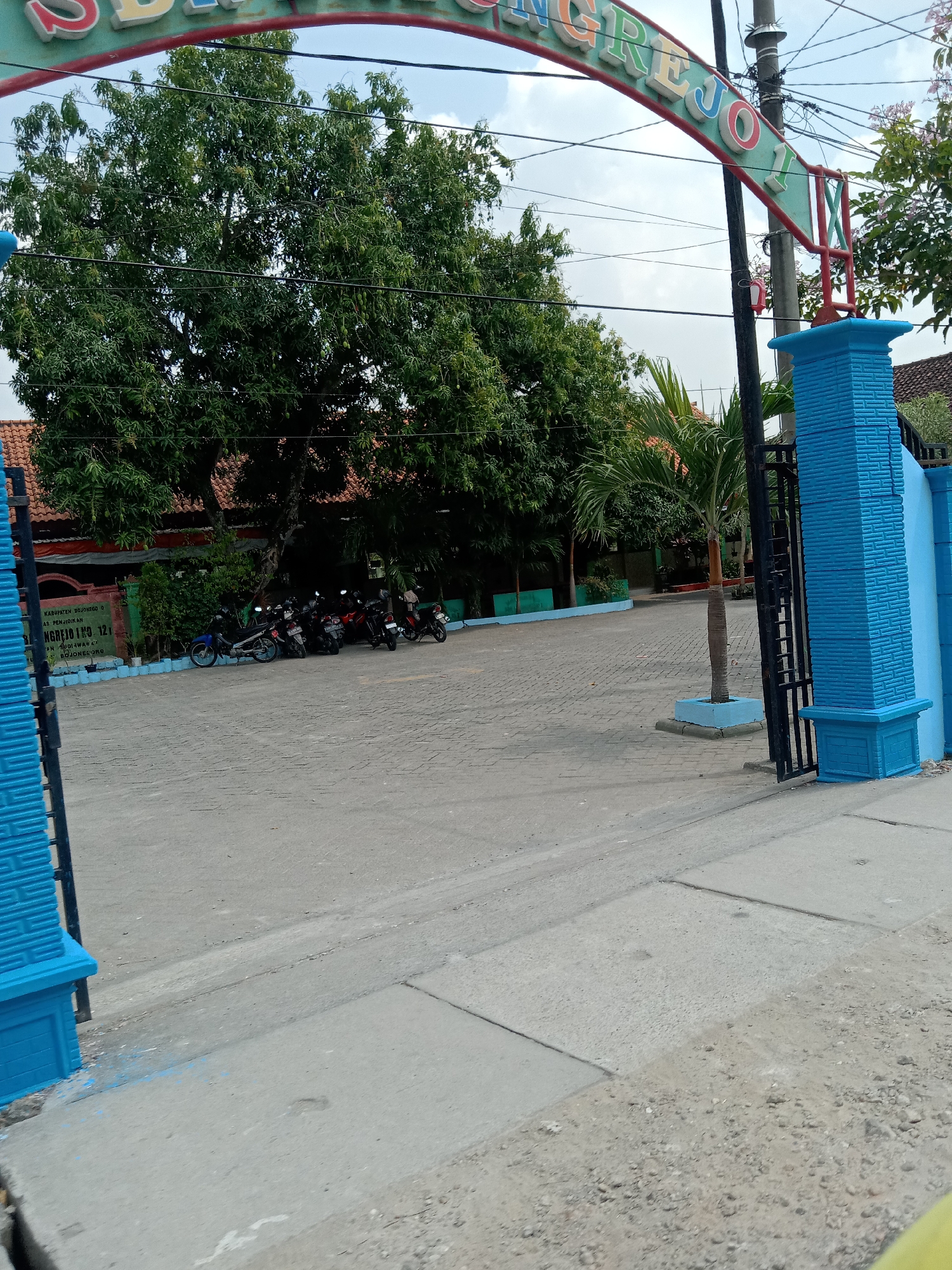 The image shows the entrance to an educational institution in Balongrejo, Indonesia. A sign above the entrance reads "SD NEGERI BALONGREJO 1," indicating it is a public elementary school. The entrance is flanked by blue brick pillars and a black metal gate. In the background, several motorcycles are parked in a courtyard. Trees and palm trees are visible, along with buildings with red tile roofs, typical of Indonesian architecture. The ground is paved with cobblestones and concrete. The sky is partly cloudy.