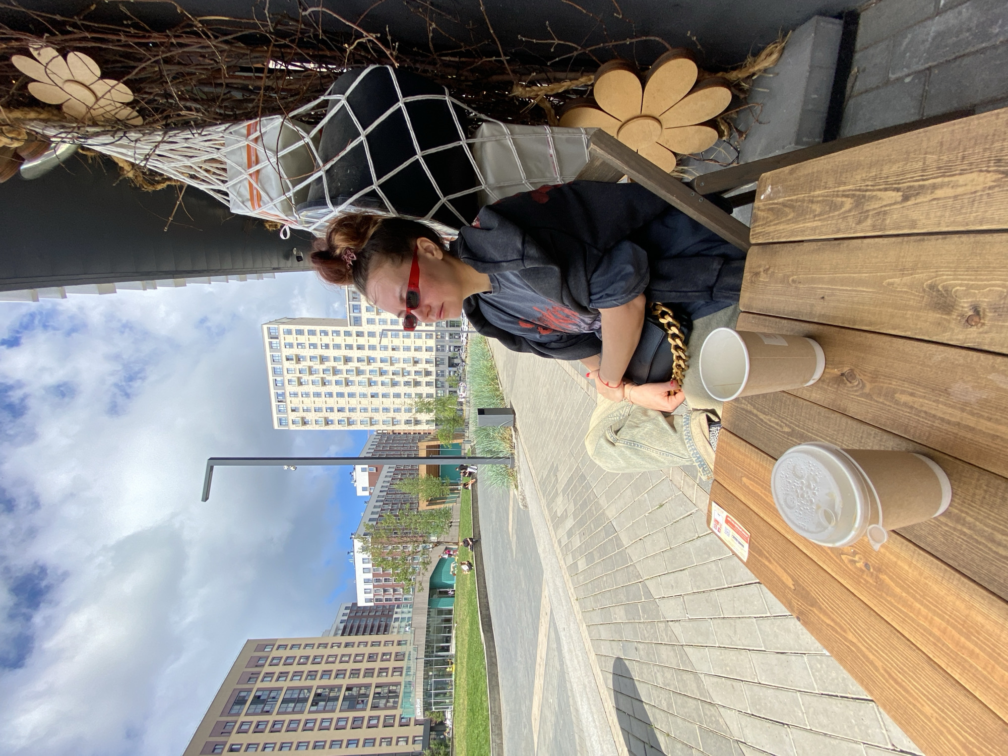 The image captures a woman sitting at a wooden table outdoors in a modern urban setting, likely in Izhevsk, Russia, as suggested by the architectural style and landscaping. The woman is wearing sunglasses and a dark t-shirt with red accents, with her hair tied up. She is seated on a chair and appears to be looking off to the side, with two paper cups placed on the table in front of her. Behind her, a decorative arrangement of branches and a wooden flower is visible, attached to a dark wall. The background reveals several contemporary apartment buildings, a paved walkway, a grassy area, and a lamppost, indicating a well-developed public space. The sky is partly cloudy.