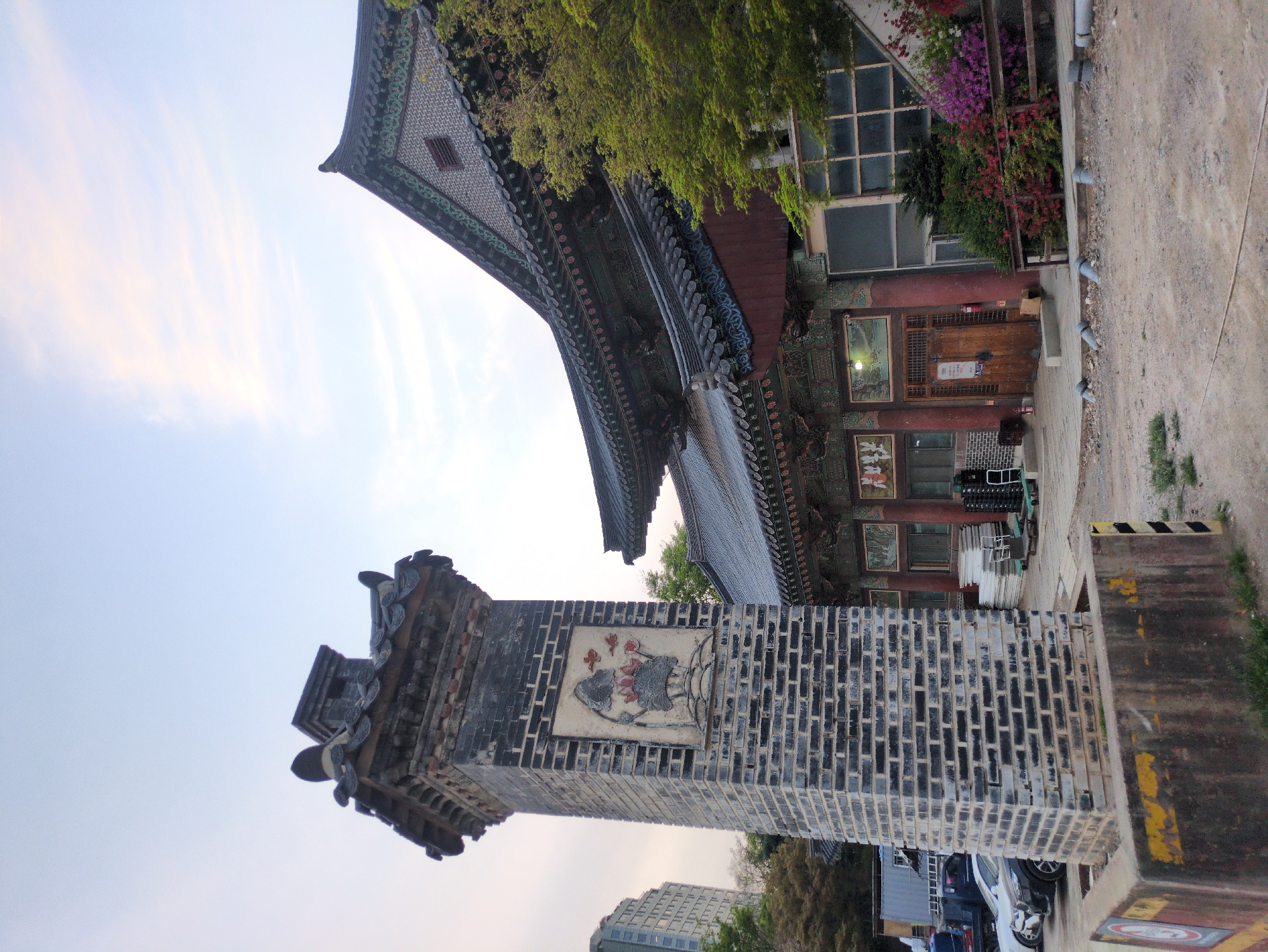 A vertically oriented photograph depicts part of a temple complex in Seoul, South Korea. The image prominently features a tall brick tower with a relief sculpture of what appears to be a lotus flower. Above and behind the tower is a traditional Korean building with a multi-tiered, ornate roof. A portion of another building with windows and potted plants is also visible to the right. The sky is partly cloudy, suggesting either early morning or late afternoon. The presence of vehicles and a modern building in the background indicates the temple is situated in an urban environment.