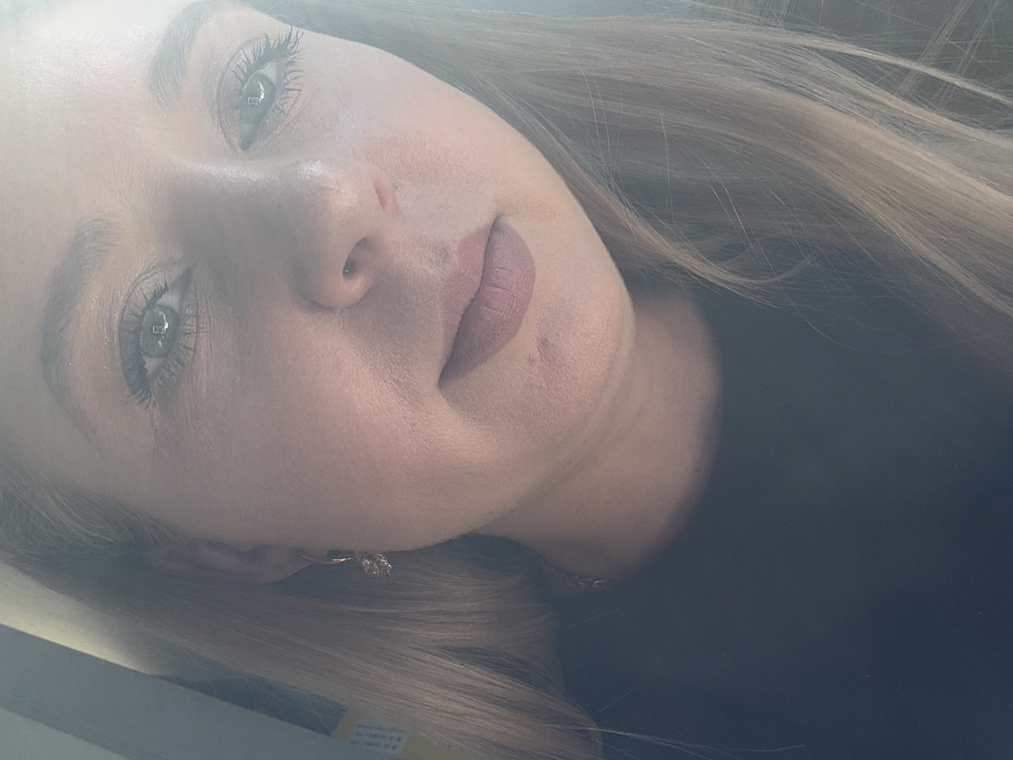 The image is a close-up, slightly overexposed selfie of a woman with blonde hair. She is looking directly at the camera. Her eyes are open, and she has on mascara. Her lips are lightly colored with lipstick. She is wearing a dark colored shirt and a dangling earring in her left ear. The background is out of focus and appears to be indoors. The lighting suggests it might be daytime, possibly near a window. The scene is a close-up of a person's face and upper chest.