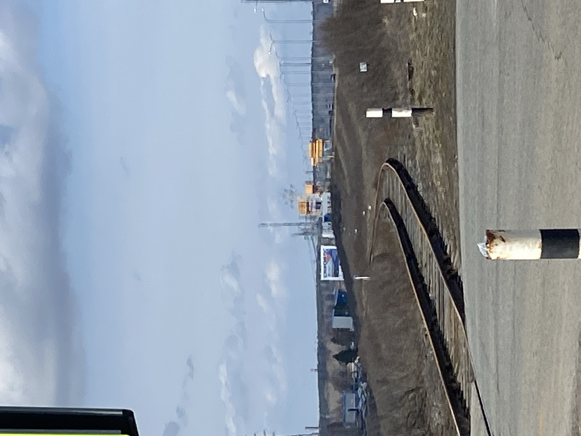 The image presents a view of the industrial landscape in Chaikovsky, Russia. Two parallel railway tracks curve into the distance, adjacent to a paved surface with safety bollards. Beyond the railway, there's a mix of bare trees, industrial buildings including a large billboard, and infrastructure such as lampposts. The sky is overcast, suggesting an overcast day. There are no people visible in the image. Yellow gantry cranes are partially visible in the distance.
