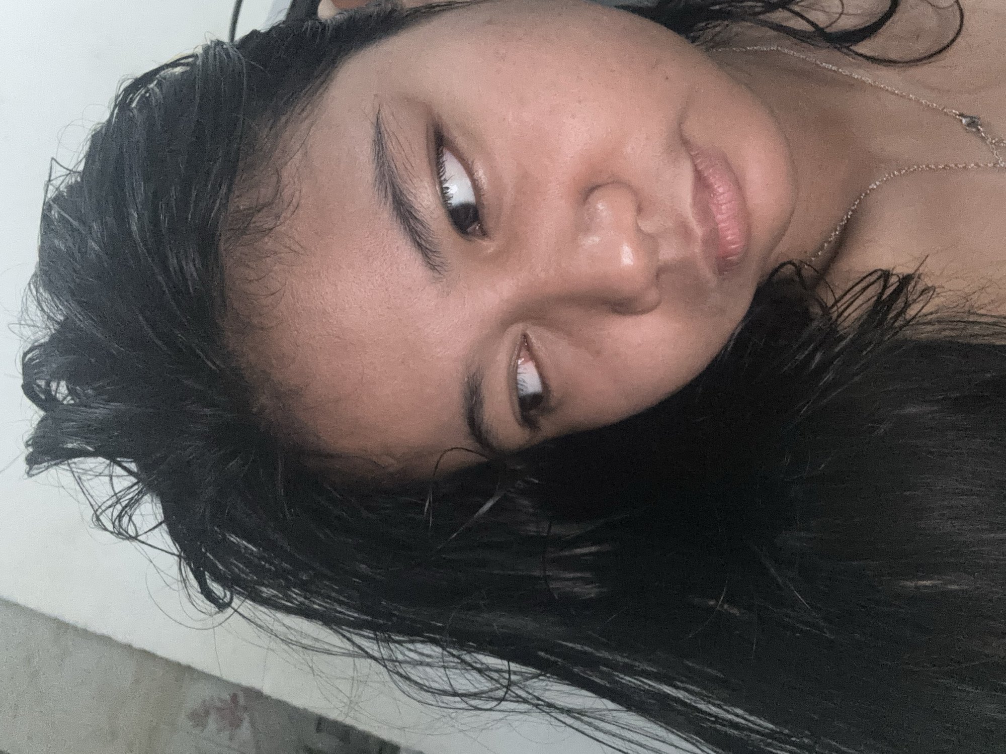 A young woman with dark, messy hair looks to her left with a neutral expression. Her skin is lightly tanned, and she appears to be indoors. The background is indistinct, with a light-colored wall and a textured surface on the left side of the frame. She is wearing a thin, silver necklace. The image is a close-up, likely a selfie, showing only her head and upper chest. There are no discernible details about the specific location within North Jakarta, Indonesia.