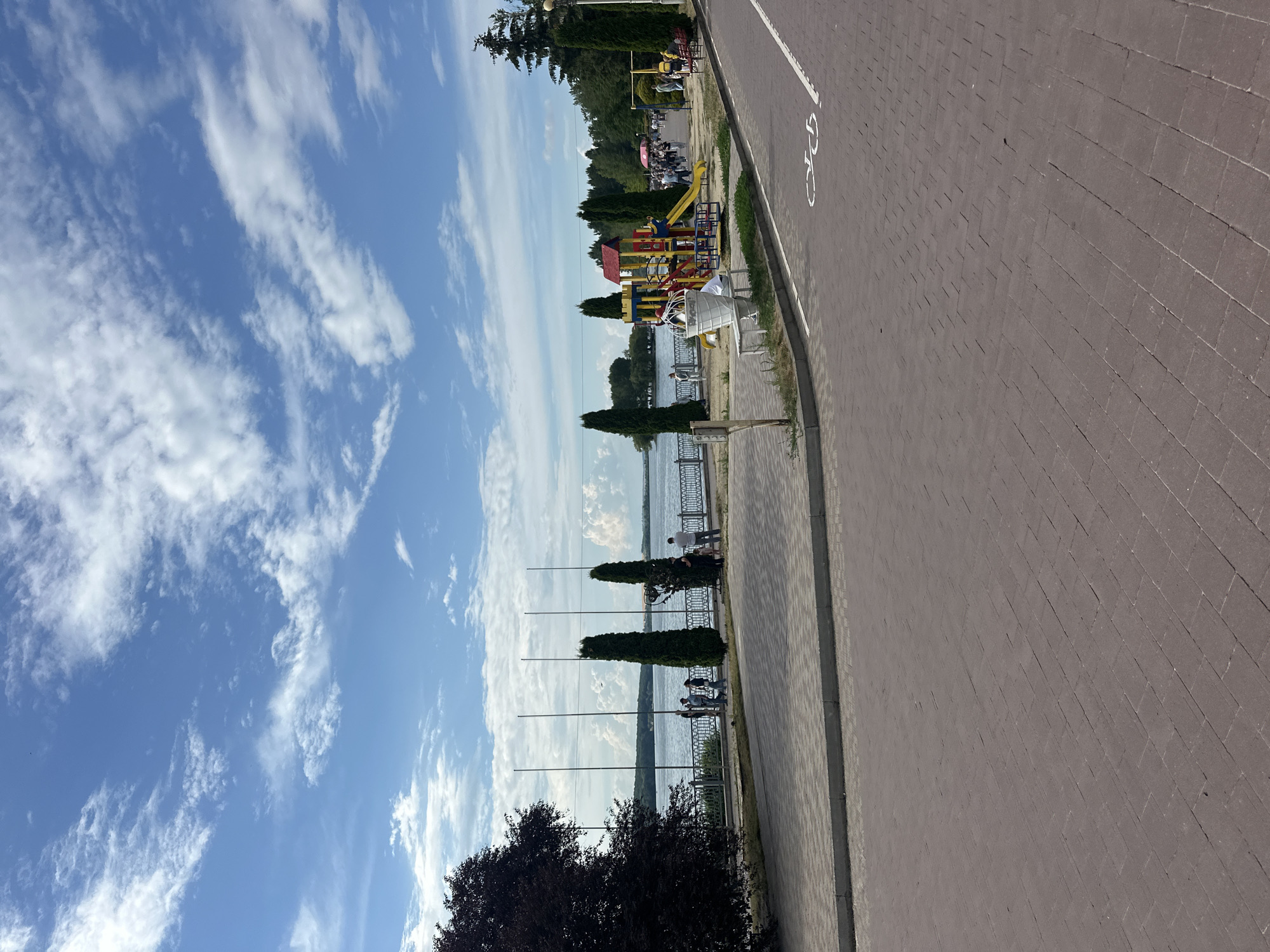 This is a sunny, daytime, eye-level outdoor photograph of a waterfront promenade with a recreational area in Ternopil, Ukraine. The foreground features a reddish-brown brick paved walkway, with a white bicycle lane marking visible on the right side.
Beyond the walkway, a paved area separates it from a grassy strip and a black metal fence that runs along the waterfront. Across the fence, a body of water, likely Ternopil Lake, stretches into the distance, meeting a treeline on the opposite shore under a bright blue sky dotted with fluffy white clouds.
To the left, a large, dark-leaved tree partially obscures the view. Along the fence line, several tall, slender green trees stand out. A few people are visible walking or standing by the water's edge, some interacting in small groups.
Further along the promenade, a colorful children's playground with slides and swings is situated next to a large decorative ice cream cone sculpture. More people are walking in this area, and a park bench is visible. The background shows a dense line of green trees. The overall atmosphere is leisurely and inviting, suggesting a public park or recreational space in the city.