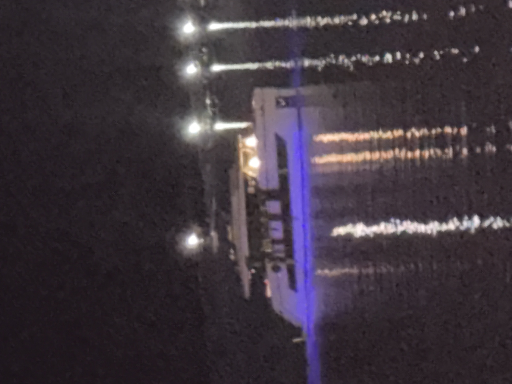 A large white yacht is illuminated with blue lights on its hull and warm yellow lights on its upper decks. The yacht is on dark water, reflecting the lights. In the background, there are several bright lights, appearing as vertical streaks on the water, suggesting they are from a distant source such as streetlights or buildings on shore. The overall scene is dark and appears to be at night. This appears to be a scene in Budva, Montenegro, possibly along the promenade or near a marina, given the presence of illuminated structures in the distance.