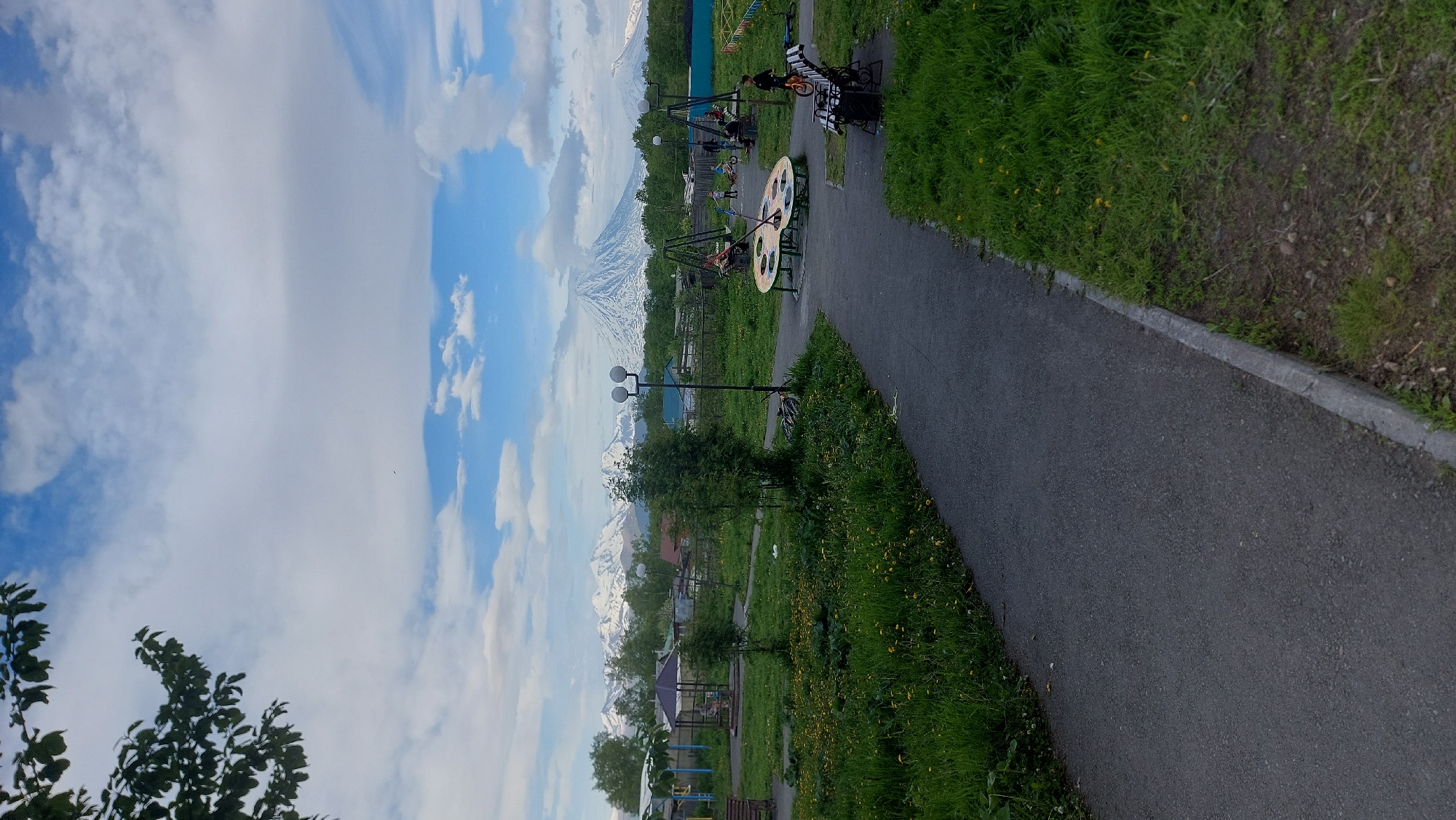 The image depicts a park scene in Zeliony, Russia, under a partly cloudy sky. The main subjects are a playground with children playing on swings and a spinning toy, and a paved walking path bordering a grassy area with scattered yellow flowers. In the background, snow-capped mountains rise majestically, partially obscured by trees and some buildings.
The activity captured is children enjoying playtime in the park. The presence of lush green grass and the bright sky suggest it is daytime, likely late spring or summer. The weather appears pleasant, with a mix of sun and clouds. The location is identifiable by the reference to Zeliony, Russia, and the distinctive mountainous backdrop.
Notable details include the playground equipment, which seems to be made of metal and wood. There are also streetlights visible, indicating the park is equipped for evening use. The path is well-maintained, suggesting a public recreational area. The overall emotion conveyed is one of peacefulness and leisure, with the natural beauty of the mountains providing a stunning backdrop to the everyday activities in the park. No visible text is present in the image.