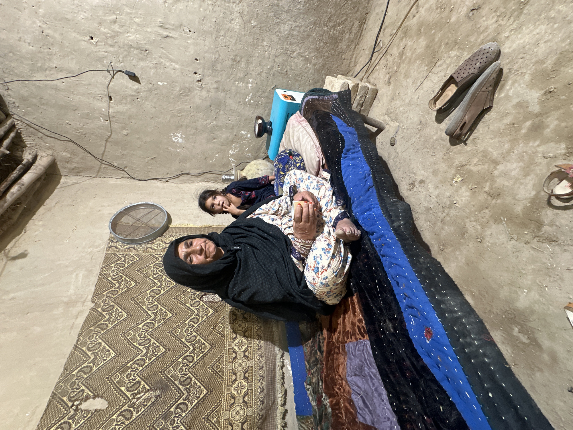 The image depicts an interior scene, presumably within a residence, located in Quetta City Tehsil, Pakistan.
An older woman wearing a dark headscarf and floral pants lies on the floor, appearing relaxed. A young girl rests near her head. They are lying on a rug. A patchwork quilt lies near them.
In the background, a blue device rests on the ground. A pair of shoes are near the wall, and a metal sieve is near the older woman. The walls and floor are textured and appear to be made of mud or plaster. A power cord is visible on the wall.