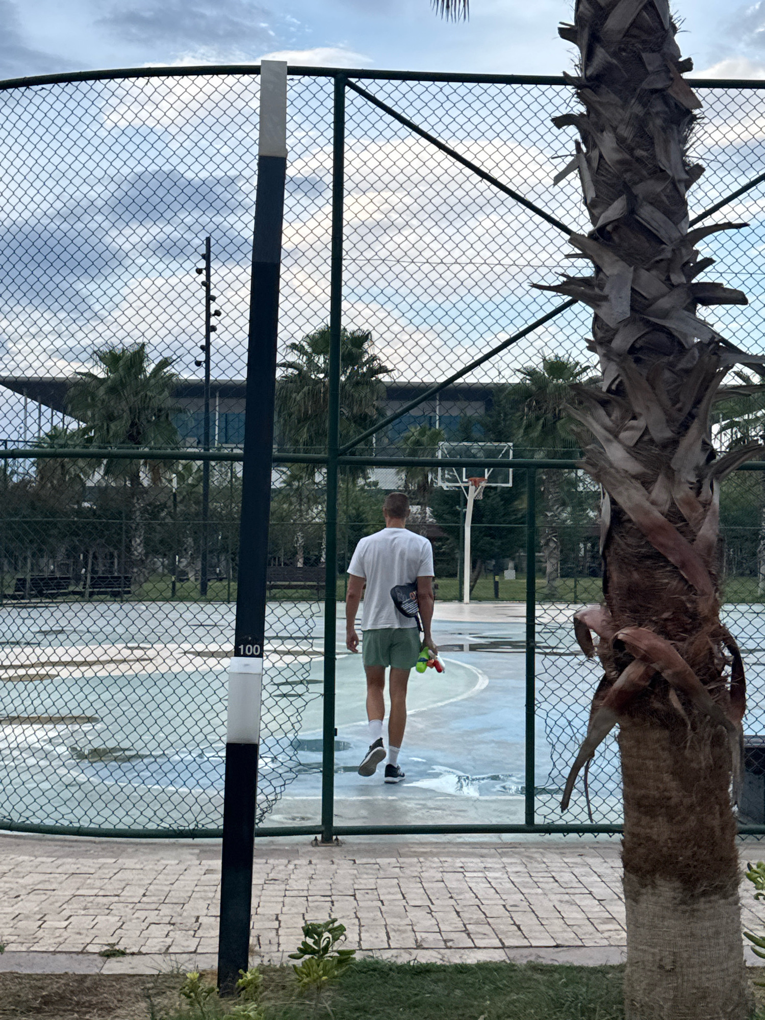 A man in a white t-shirt and green shorts walks away from the camera on a wet basketball court, holding two small water guns. He is positioned in the center of the frame, walking towards a basketball hoop visible in the background. He is wearing black athletic shoes and white socks. The court appears to be outdoors, with a chain-link fence enclosing it. Several palm trees are visible around the court, with one prominently in the foreground on the right side of the image. In the background, a modern building with large windows can be seen behind the palm trees. The sky is cloudy. A black and white striped pole is present on the left side of the image, with the number "100" visible on a white section near its base. The scene suggests a leisure activity taking place at an outdoor sports facility, potentially in a resort or recreational area. The image was captured in Antalya, Turkey.